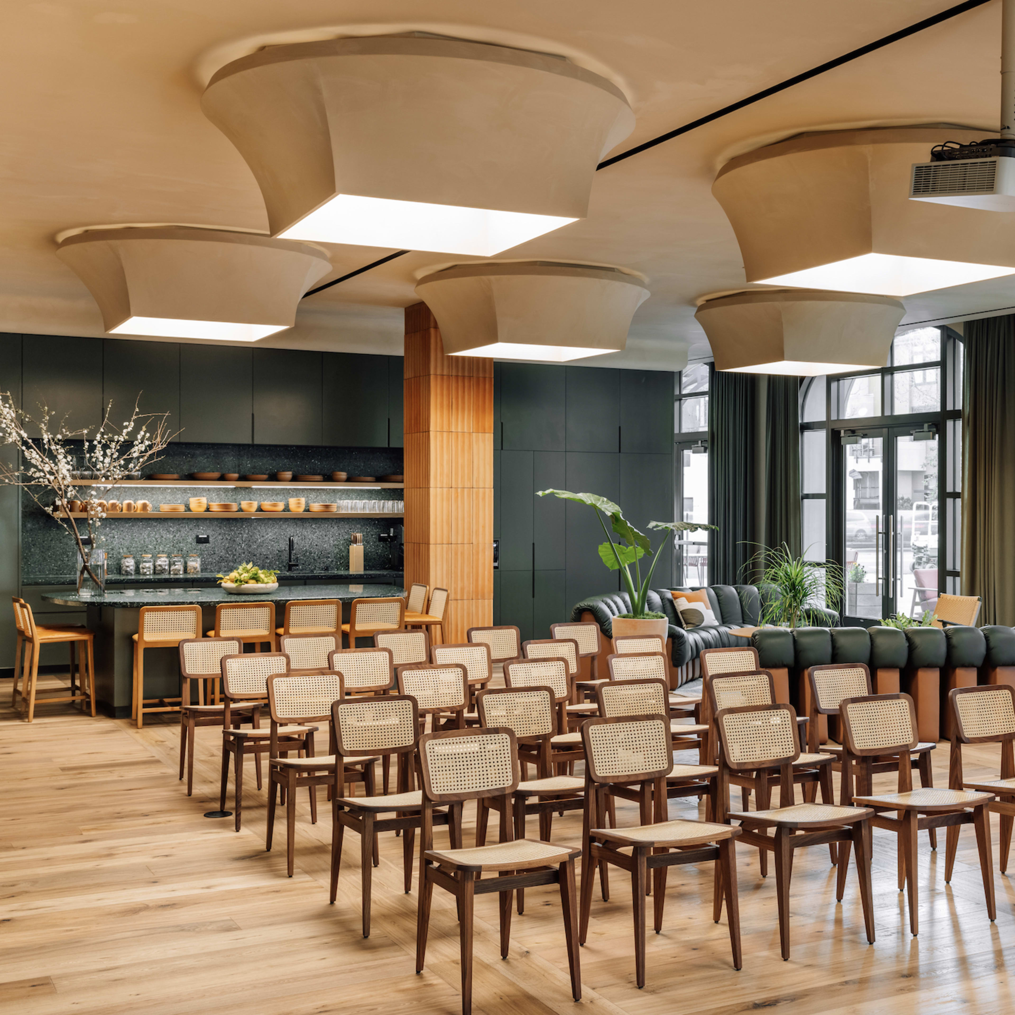 A modern interior space with rows of wooden chairs arranged facing a dark kitchen area, featuring large light fixtures and a mix of seating areas.