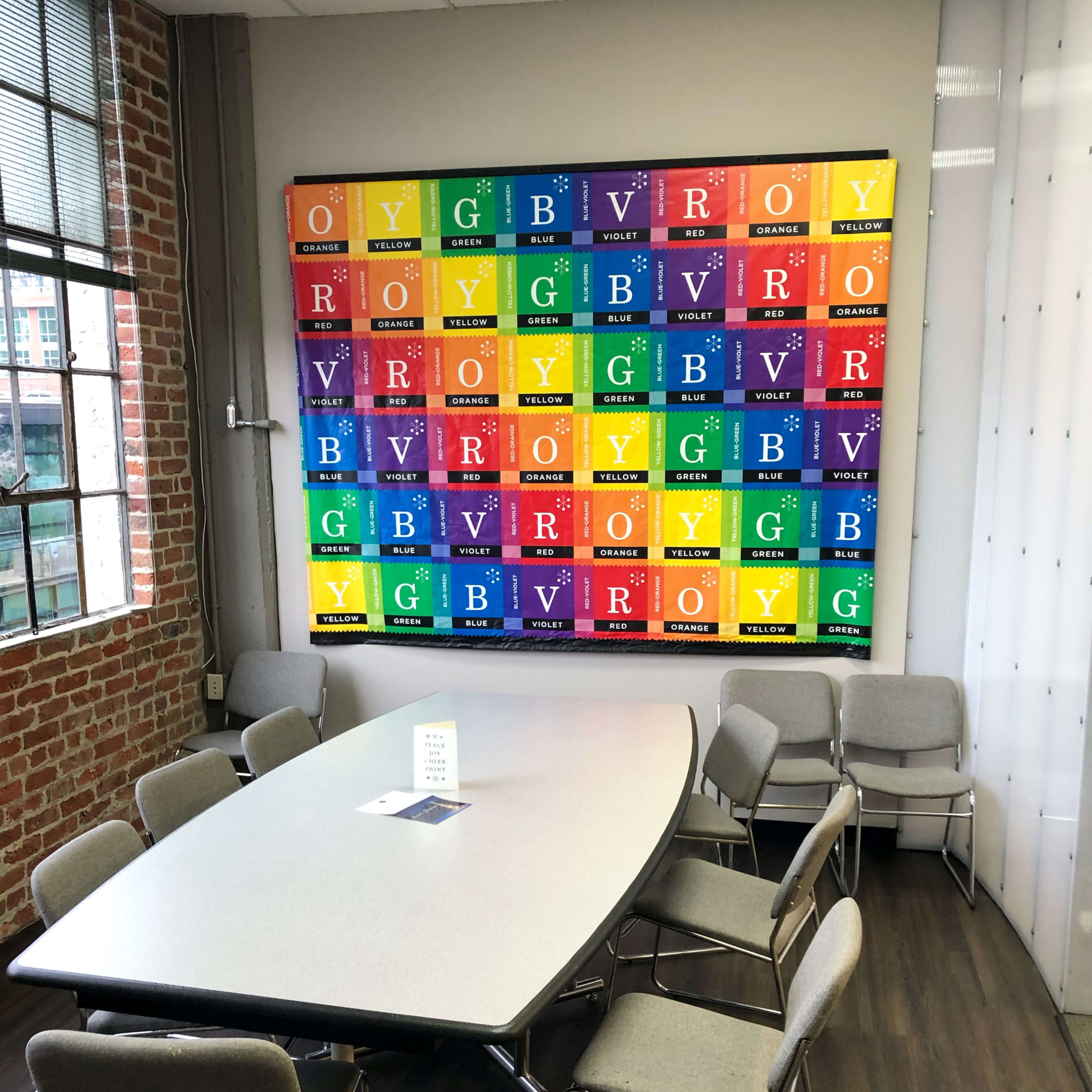 A meeting room with a rectangular table surrounded by chairs, featuring a colorful periodic table artwork mounted on the wall.