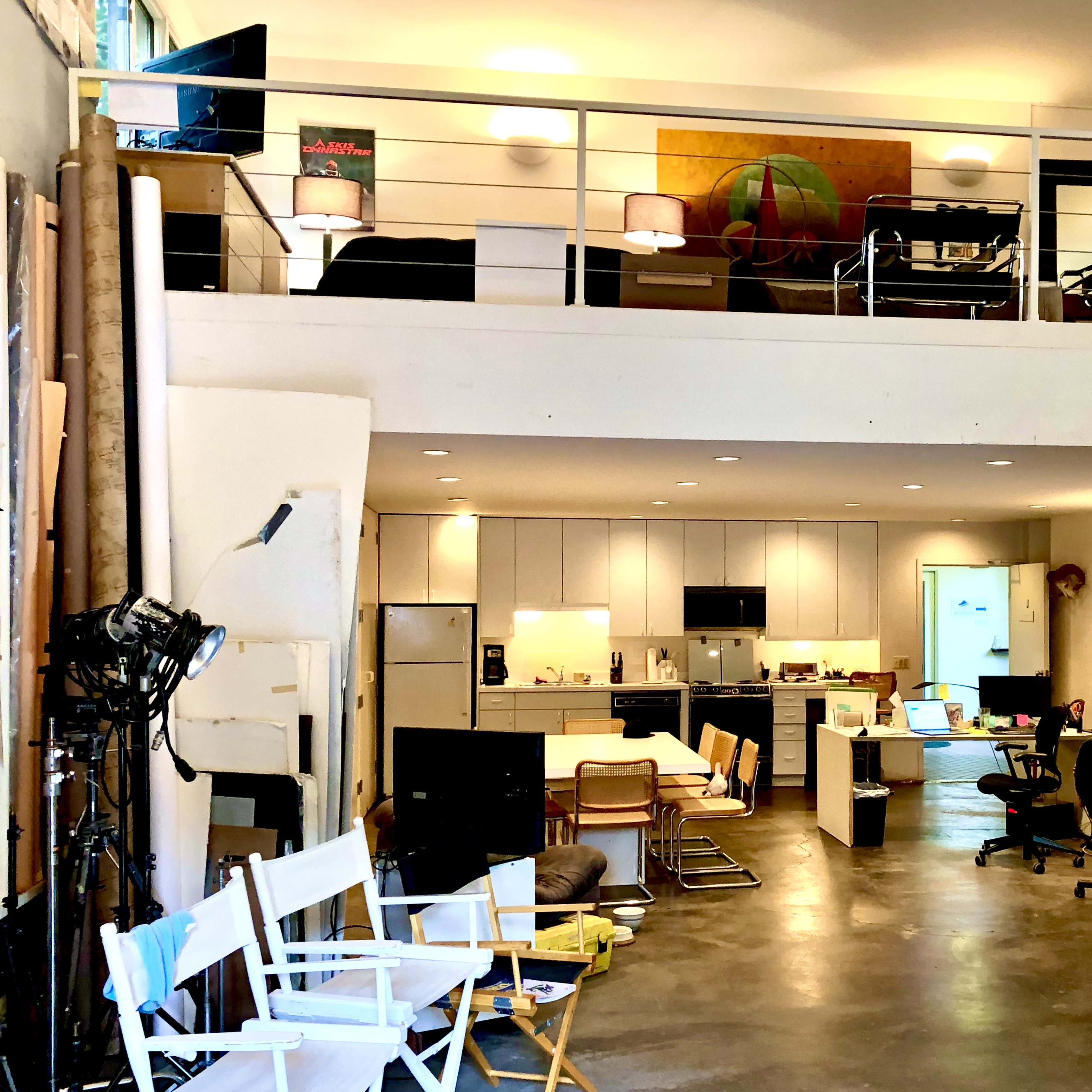 A spacious, multi-level studio apartment featuring a kitchen, workstations, and a loft area with a sitting space.