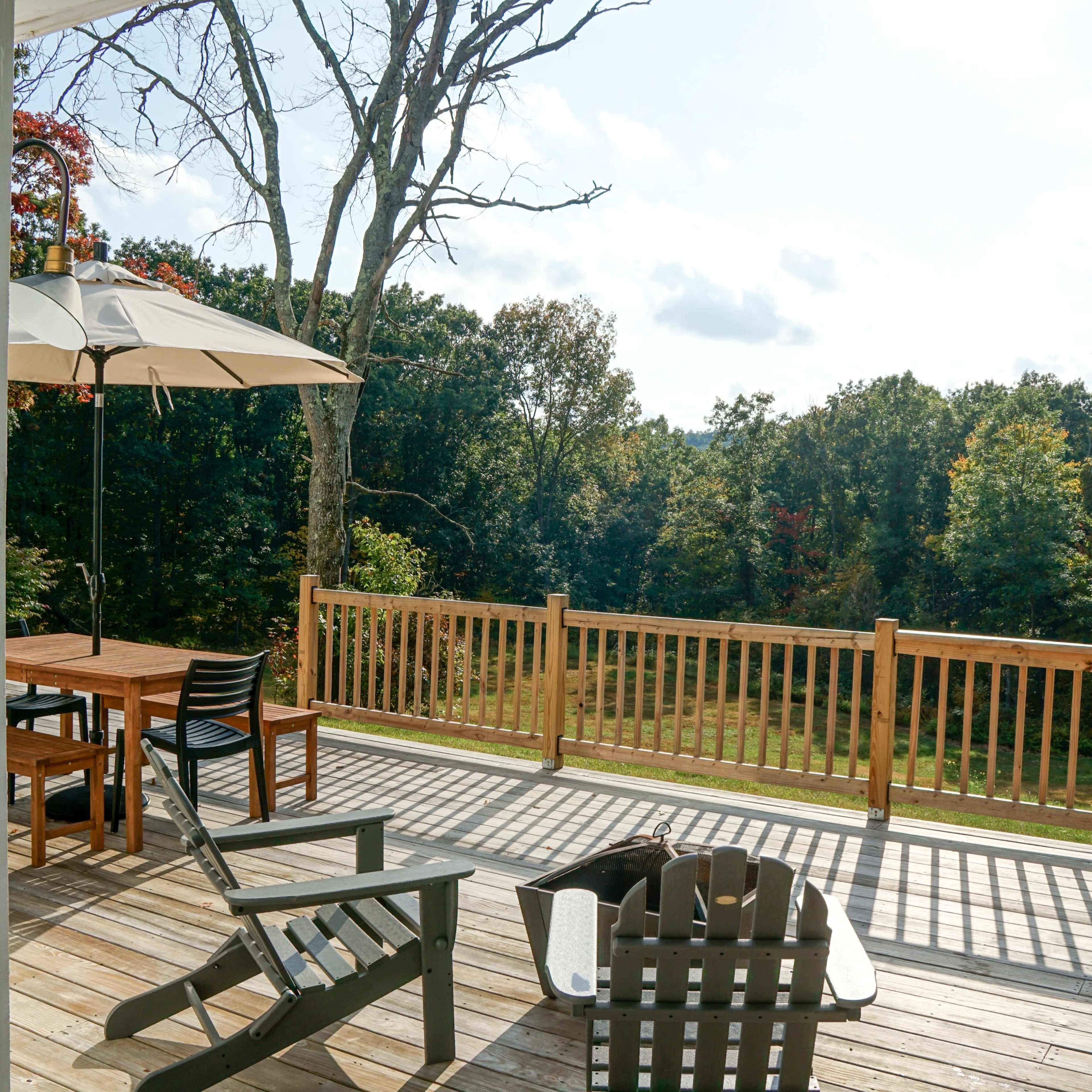 A wooden deck features a dining table, chairs, and an umbrella, overlooking a grassy area surrounded by trees.