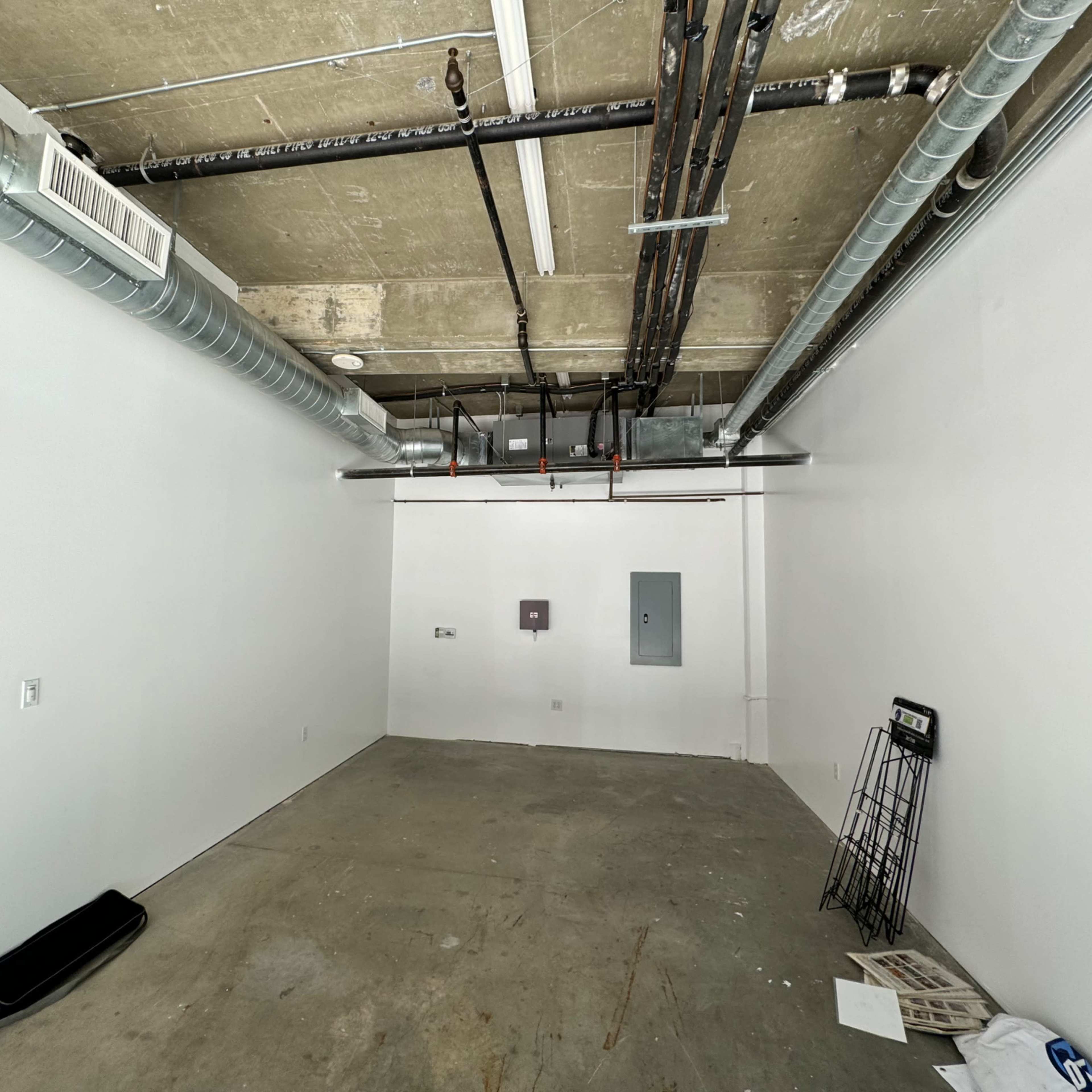 An empty room with bare concrete flooring, white walls, exposed ductwork, and a few electrical outlets.