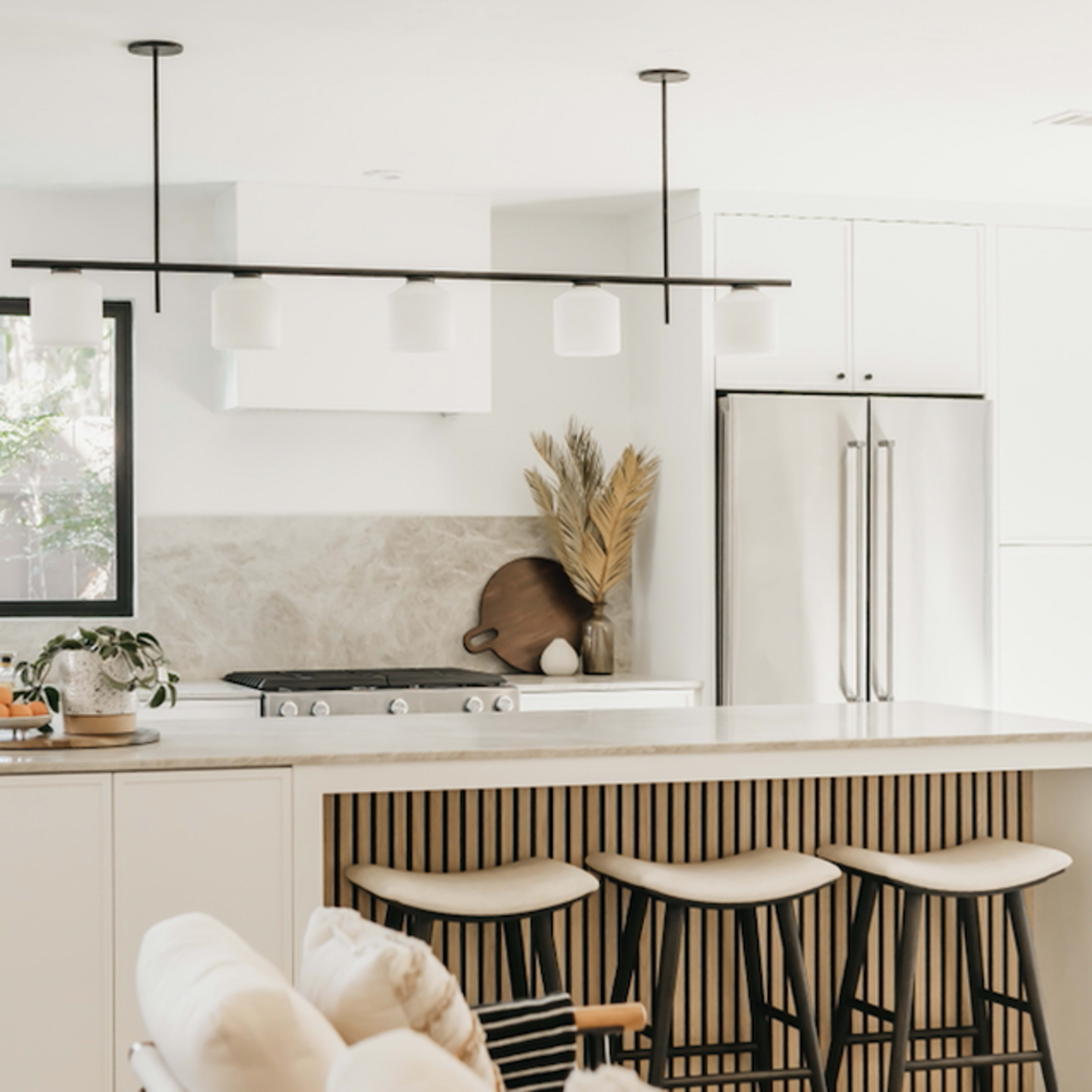 A modern kitchen features a central island with bar stools, white cabinetry, and a large window allowing natural light to illuminate the space.