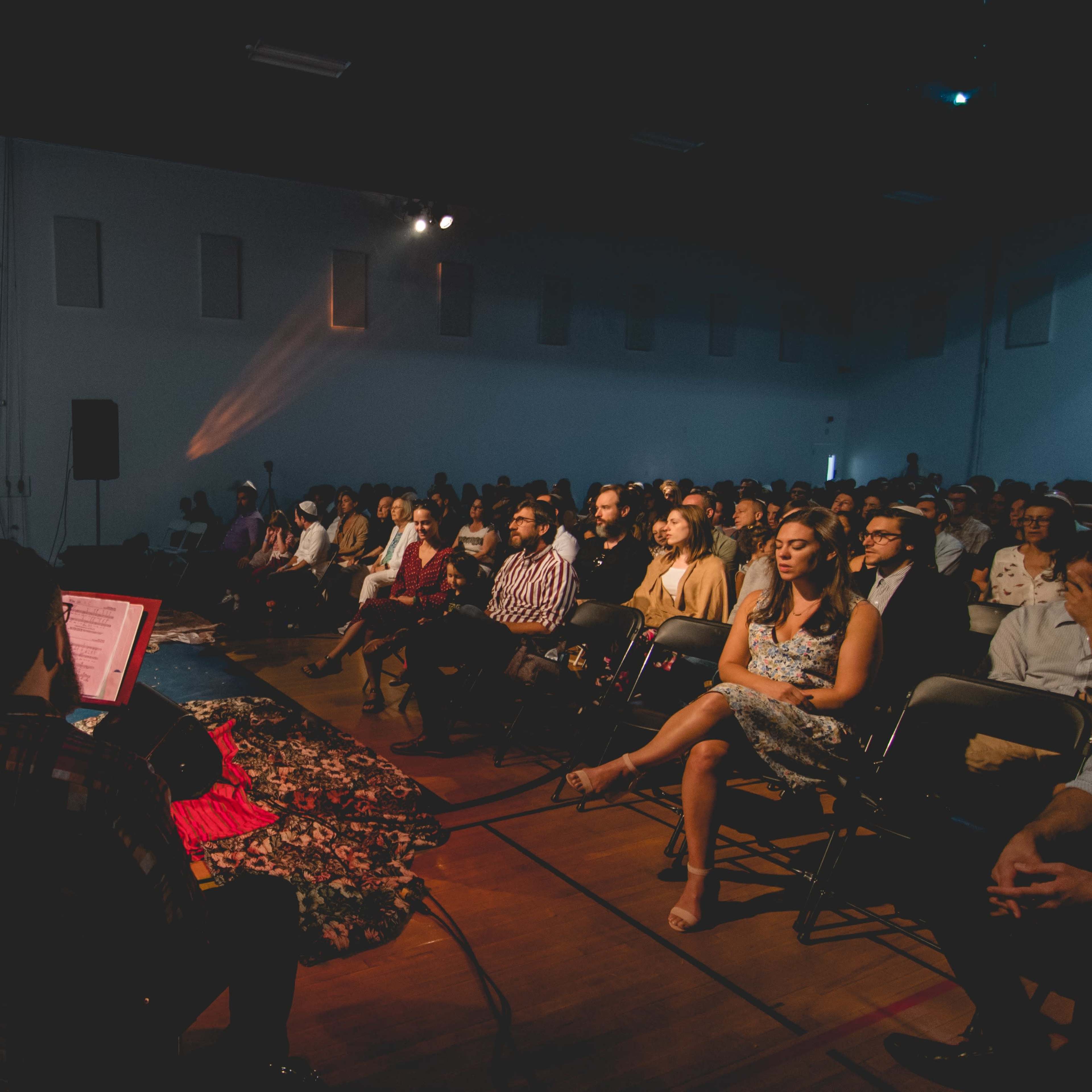 A group of musicians performs on stage in front of an audience seated in a spacious indoor venue.