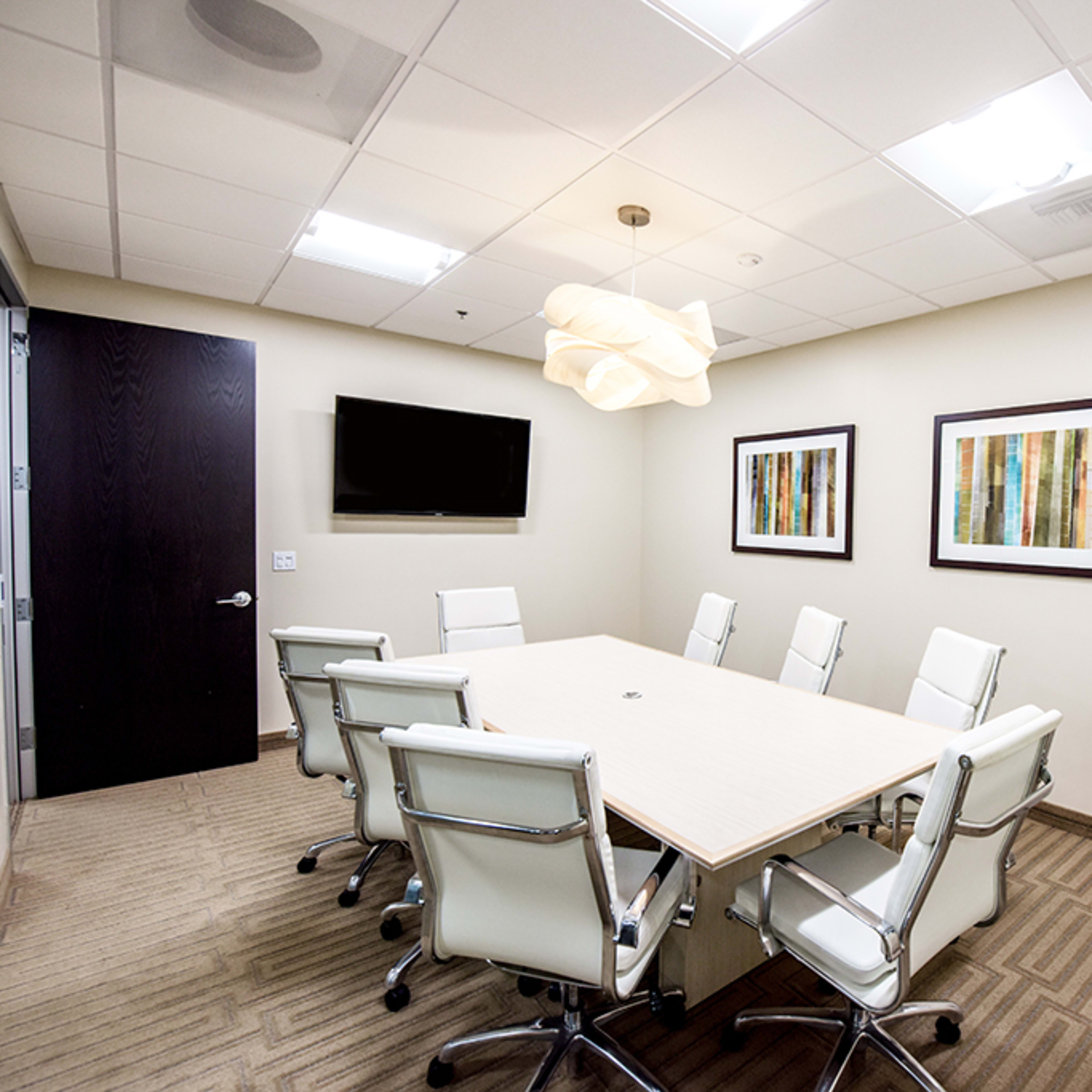A modern conference room features a large rectangular table surrounded by white leather chairs, a mounted television, and two framed artworks on the light-colored walls.