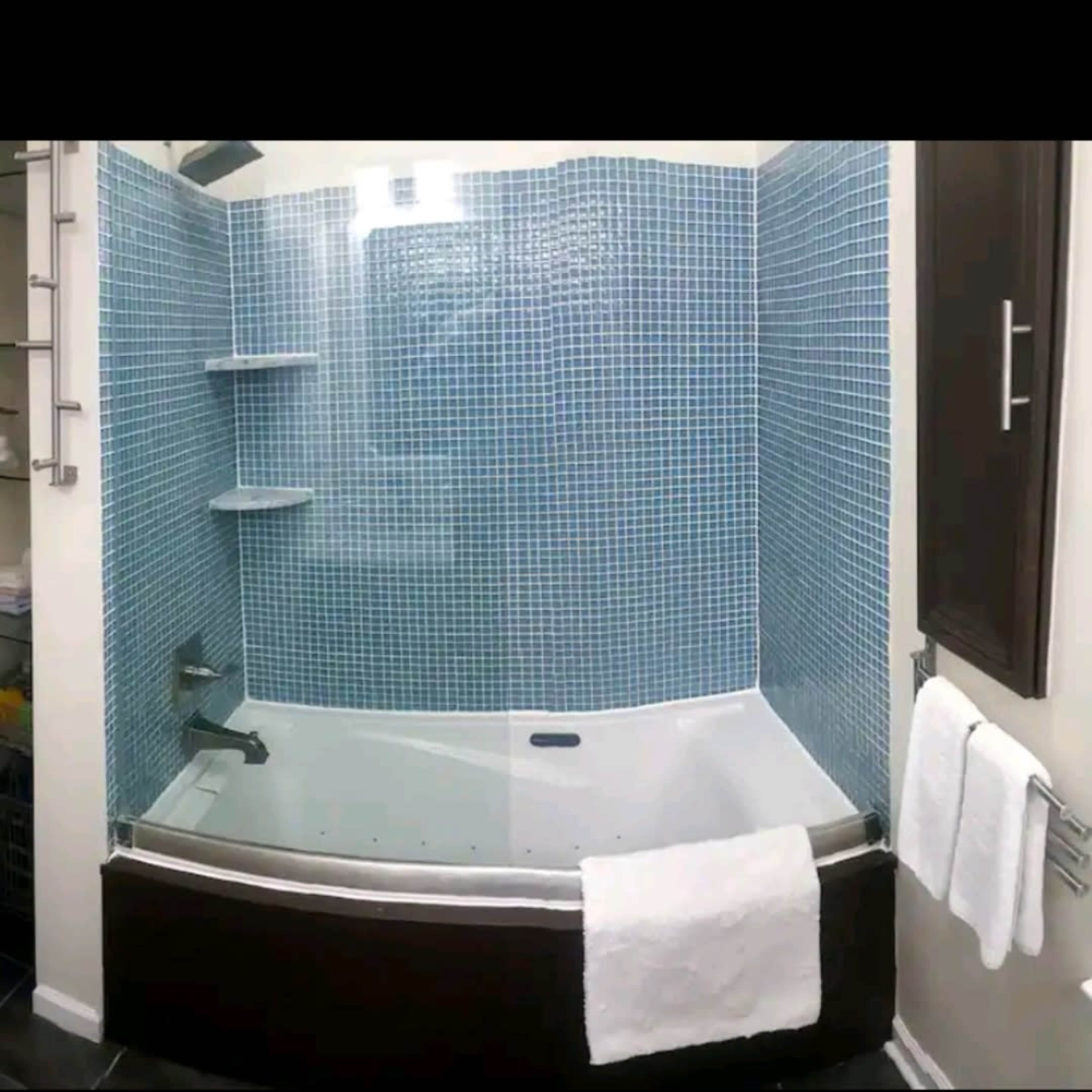 A modern bathroom with a curved glass shower enclosure, a blue tiled wall, and a white bathtub surrounded by shelves and towels.