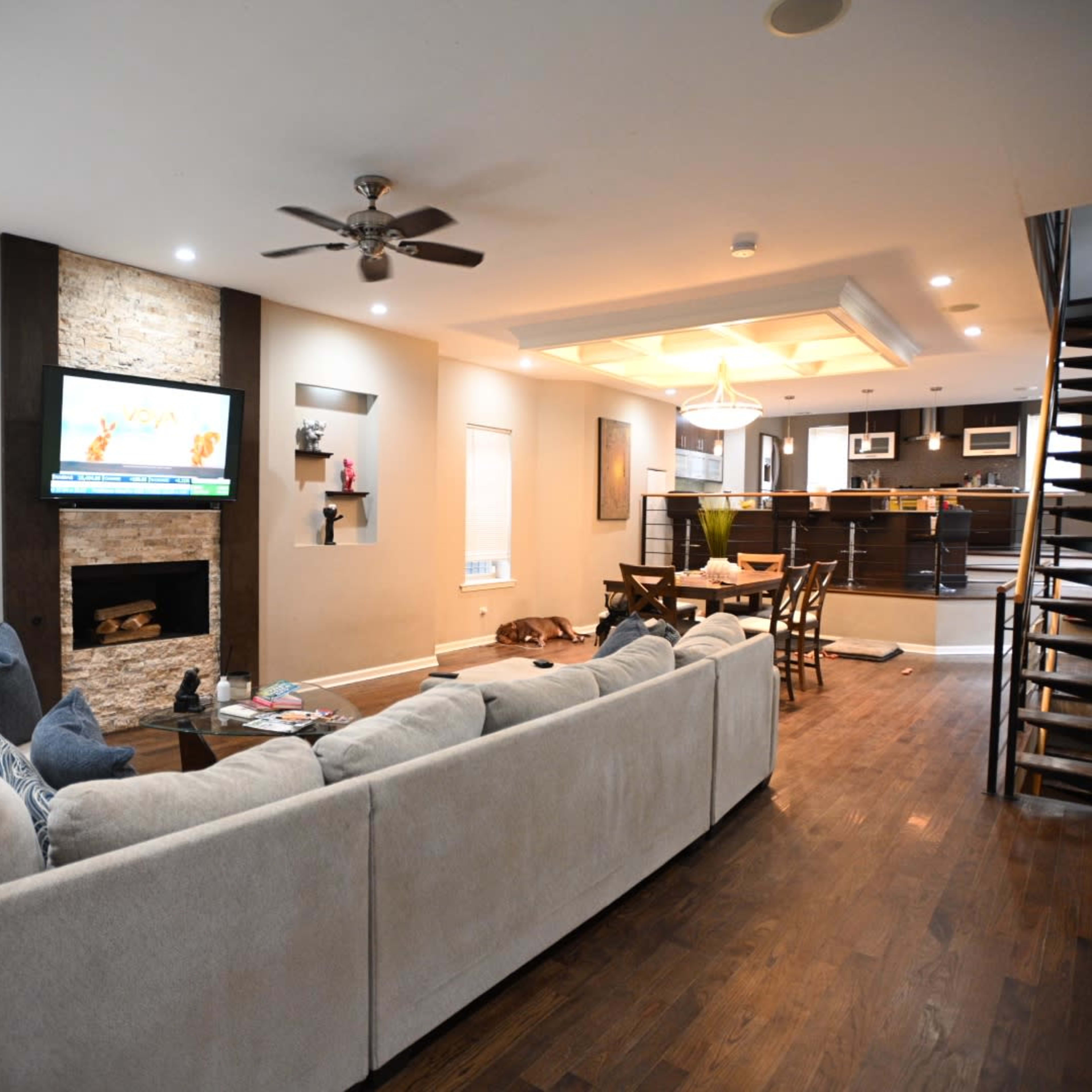 A modern living room with a beige sectional couch, a stone fireplace with a TV above it, and a staircase leading to an upper level.