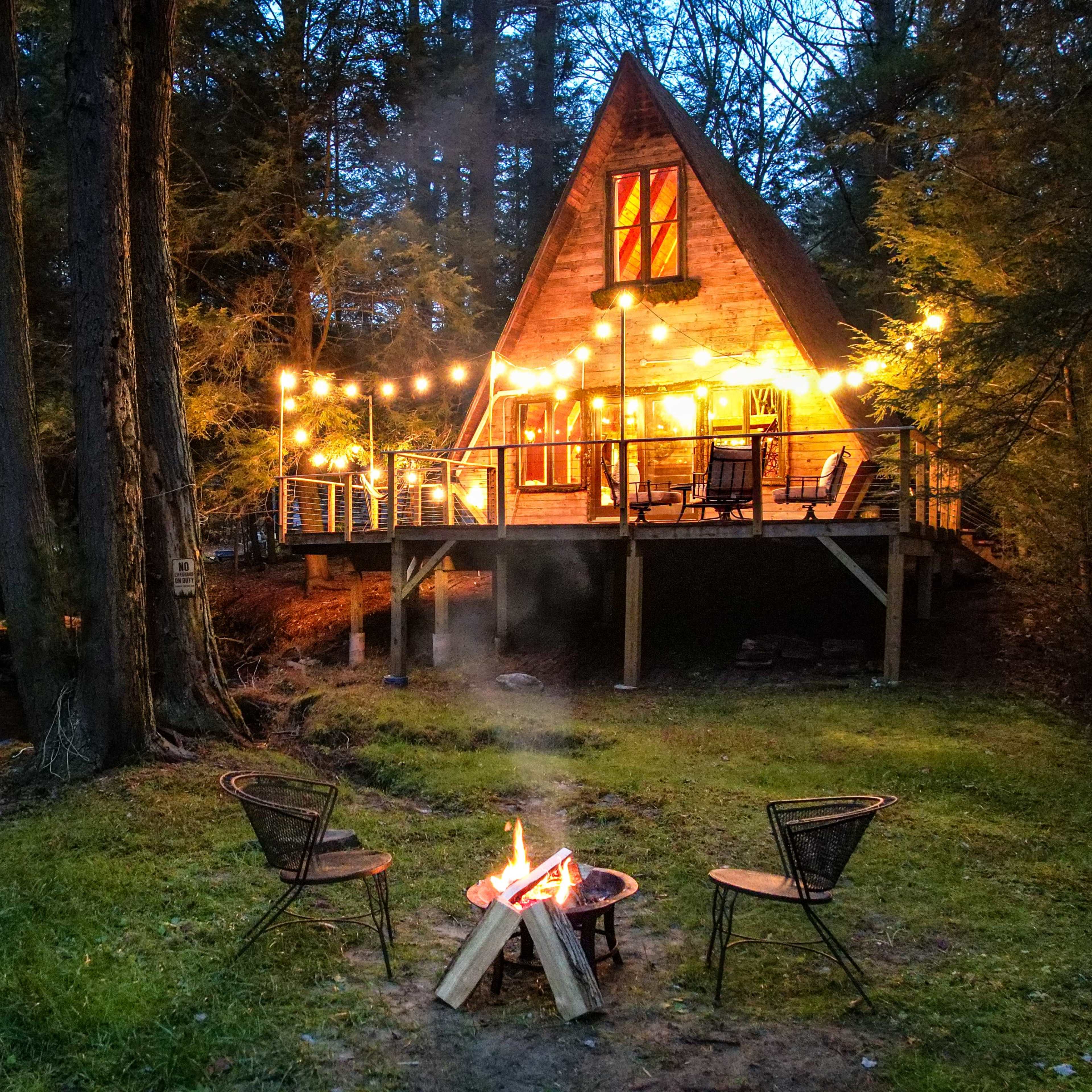 A lit A-frame cabin stands among trees, with a fire pit and three chairs arranged in front of it near a flowing stream.