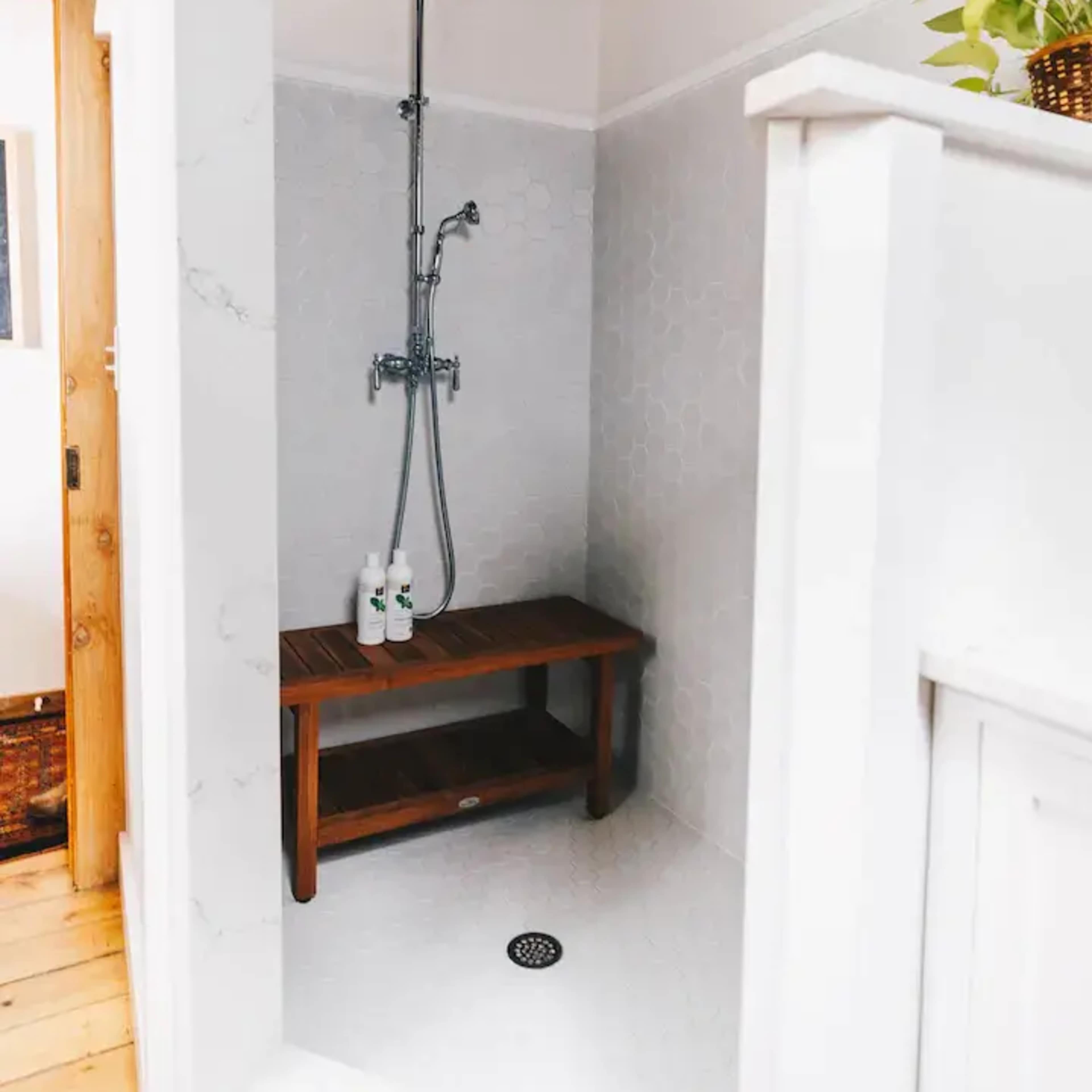 A corner shower with a wooden bench, a wall-mounted showerhead, and a tiled floor.