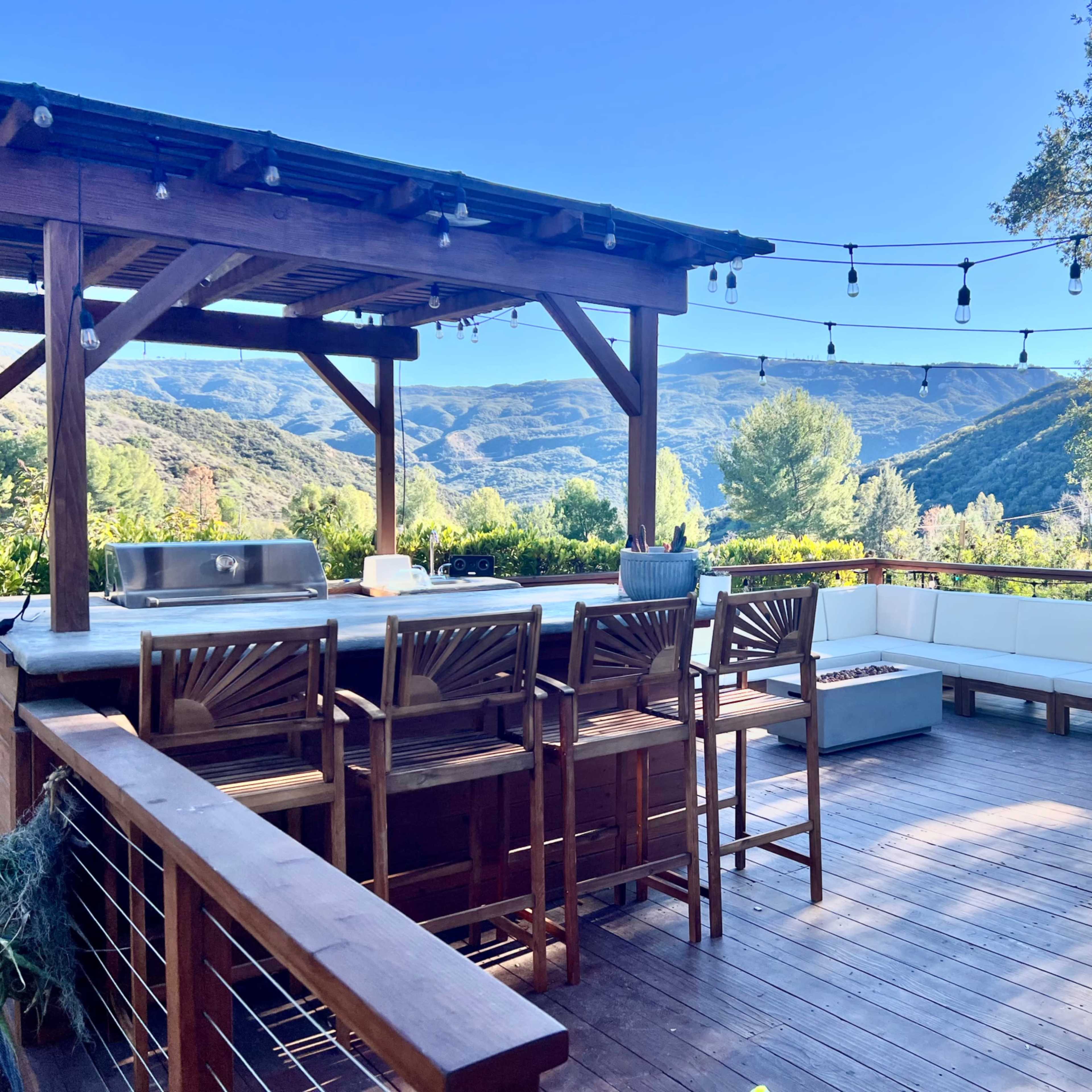 A garden area with metal planters, a greenhouse, and a house under a bright sun against a backdrop of blue skies and mountains.
