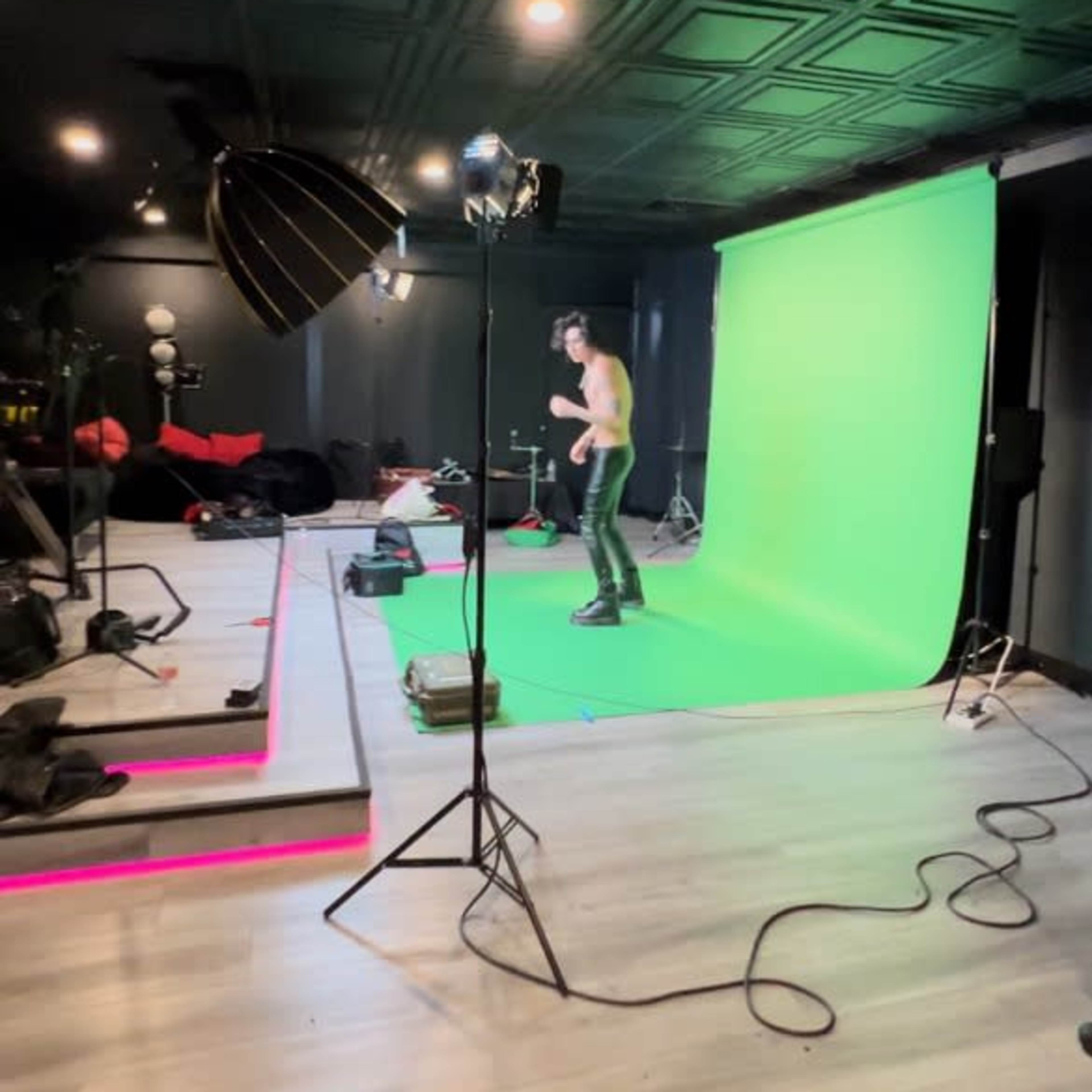 A person poses in front of a green screen in a studio setting equipped with lighting and various equipment.