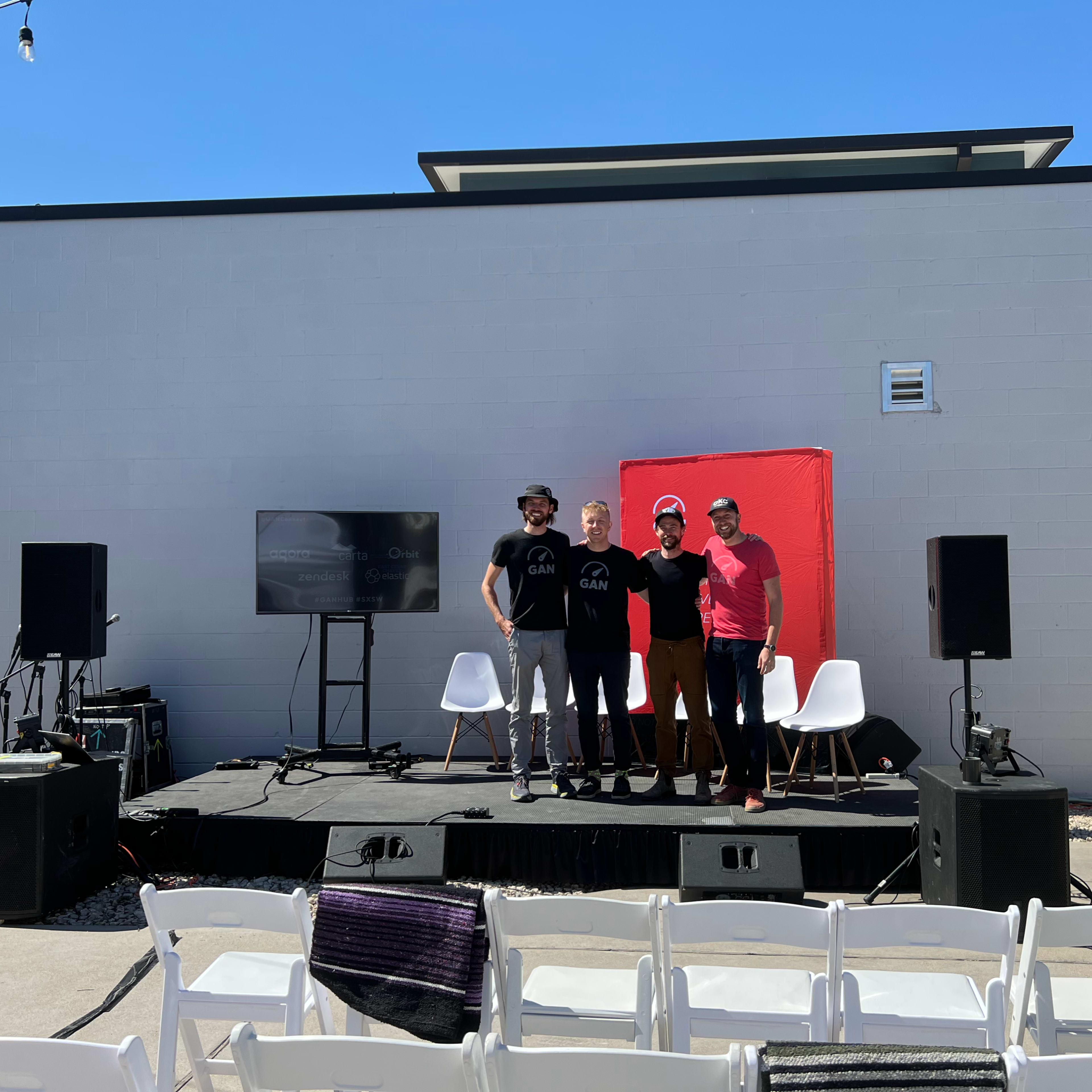 A group of four men stands together on a stage in front of a blank wall, with chairs and audio equipment arranged for an event.