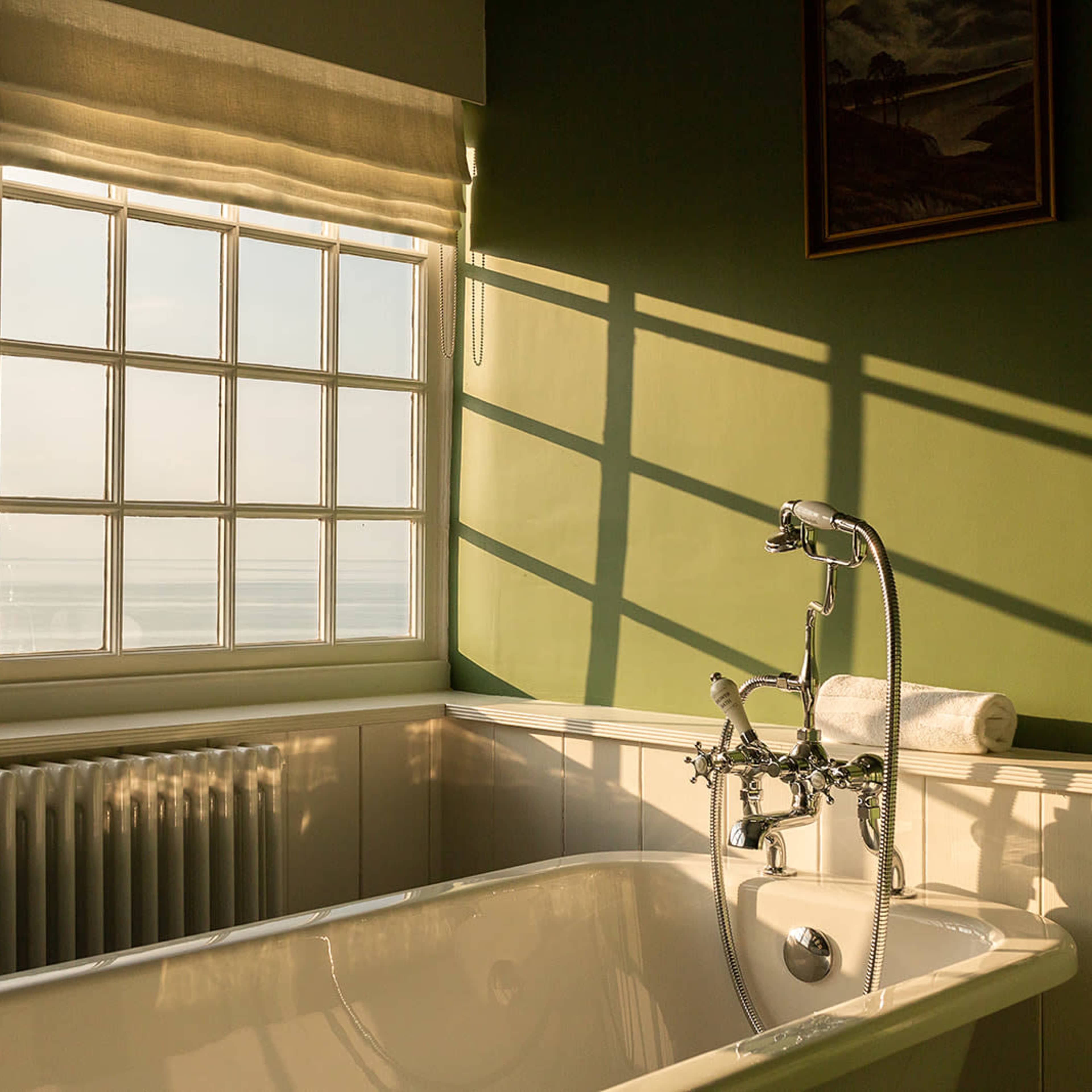 A bathtub is positioned next to a large window, with sunlight casting shadows on the green wall and a view of the sea in the background.