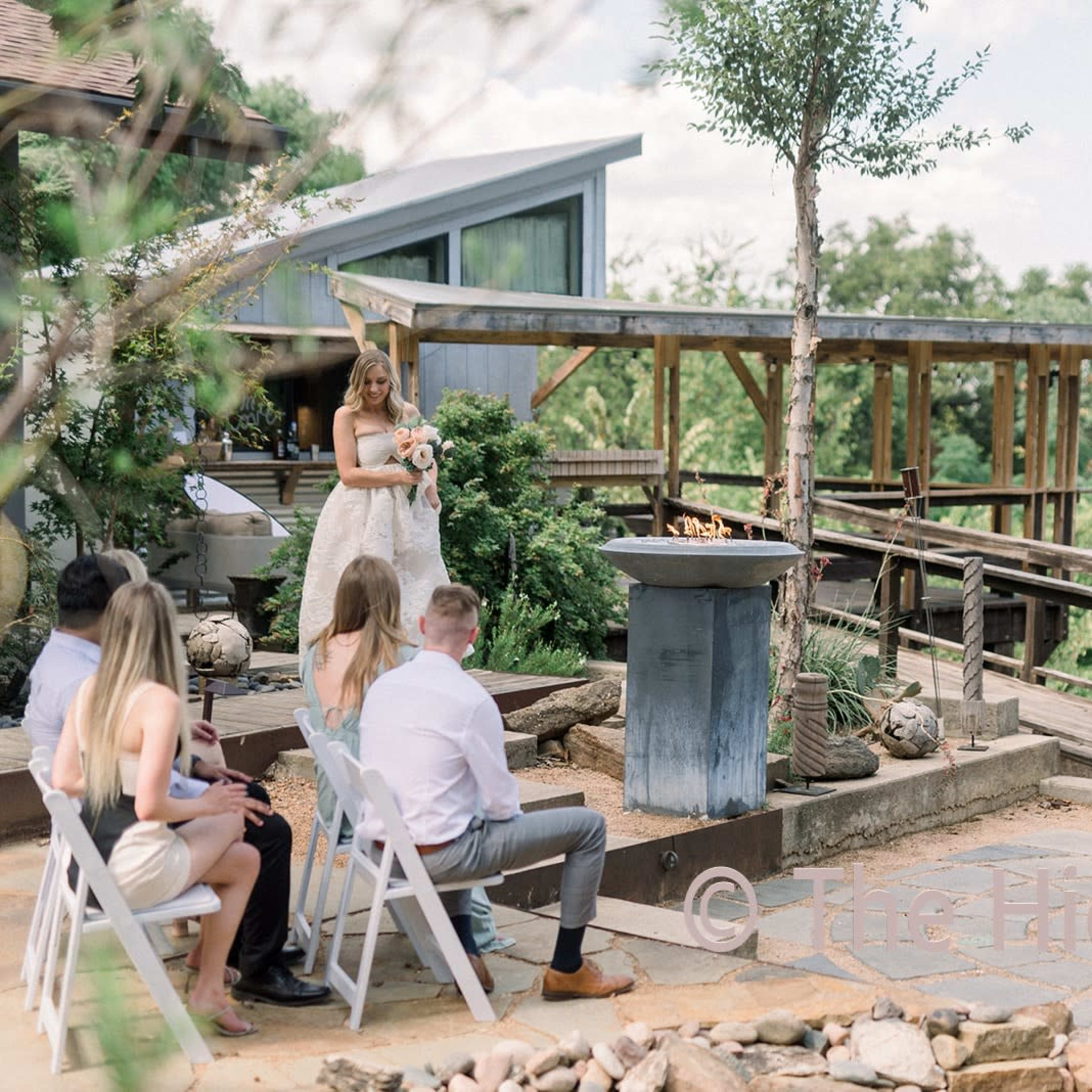 A bride stands with a bouquet in a garden ceremony while a groom faces her, surrounded by guests seated on white chairs.