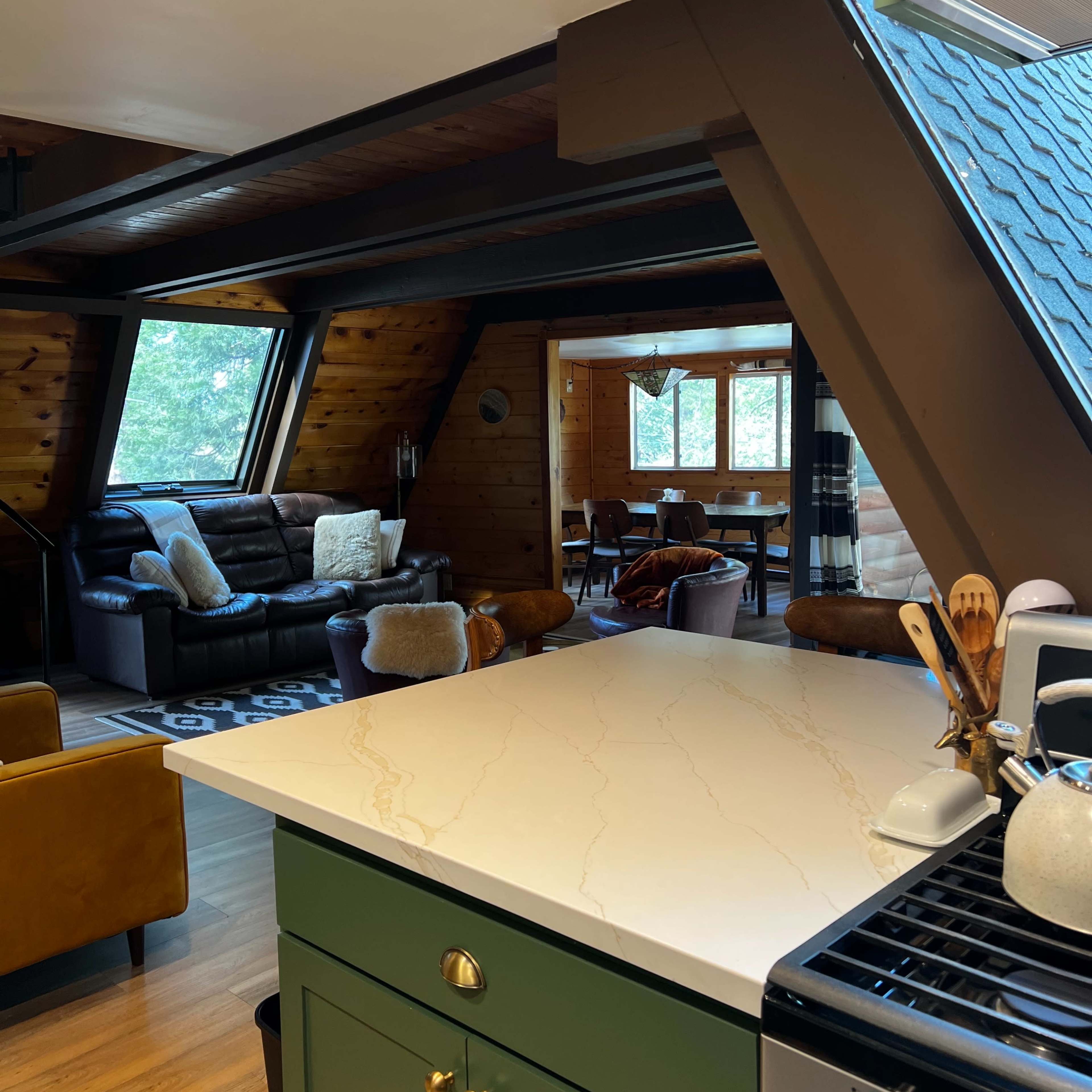 A modern kitchen with a green cabinet and marble countertop, connected to a cozy living area featuring a leather sofa and a dining space in an A-frame style home.
