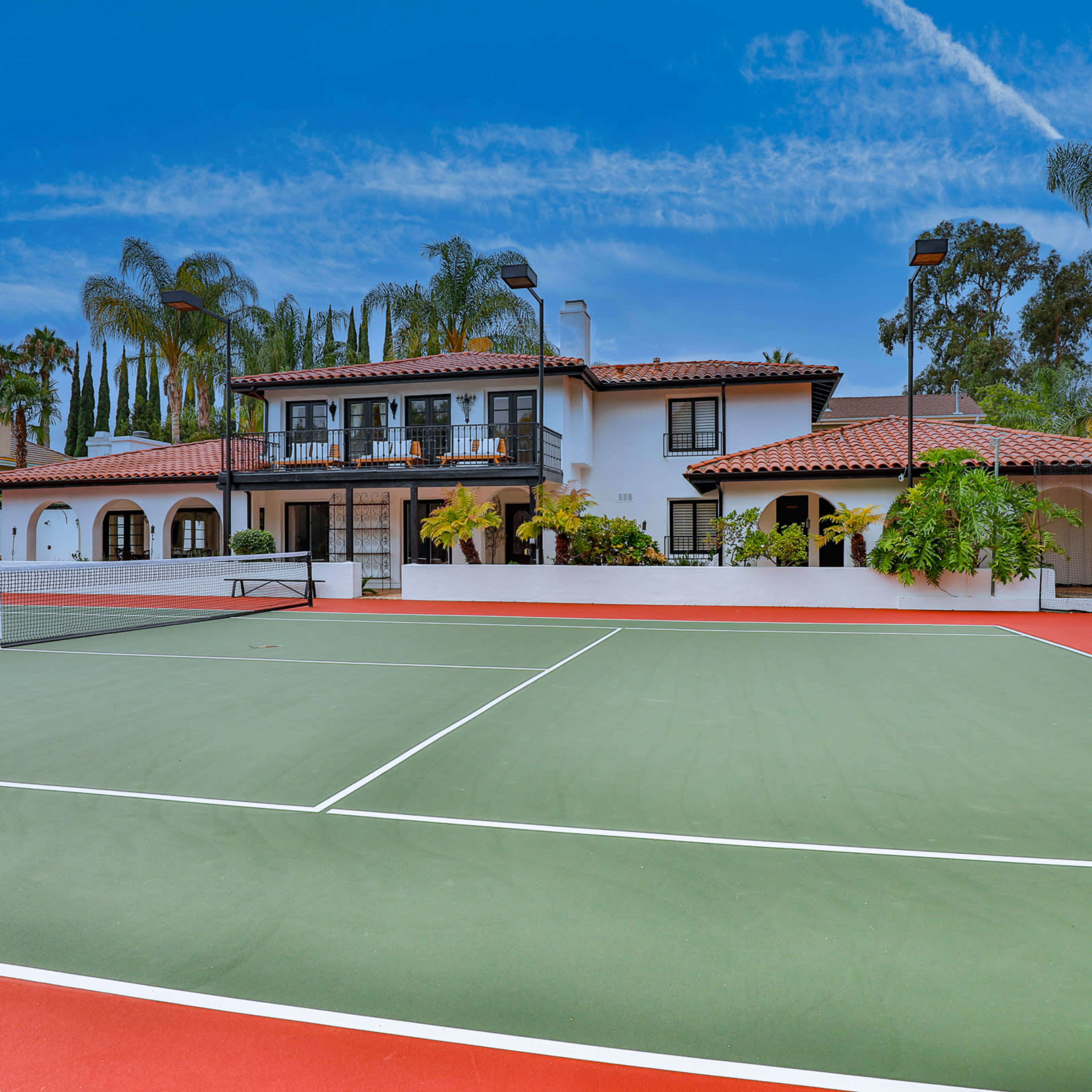 A tennis court in front of a two-story house surrounded by palm trees and greenery.