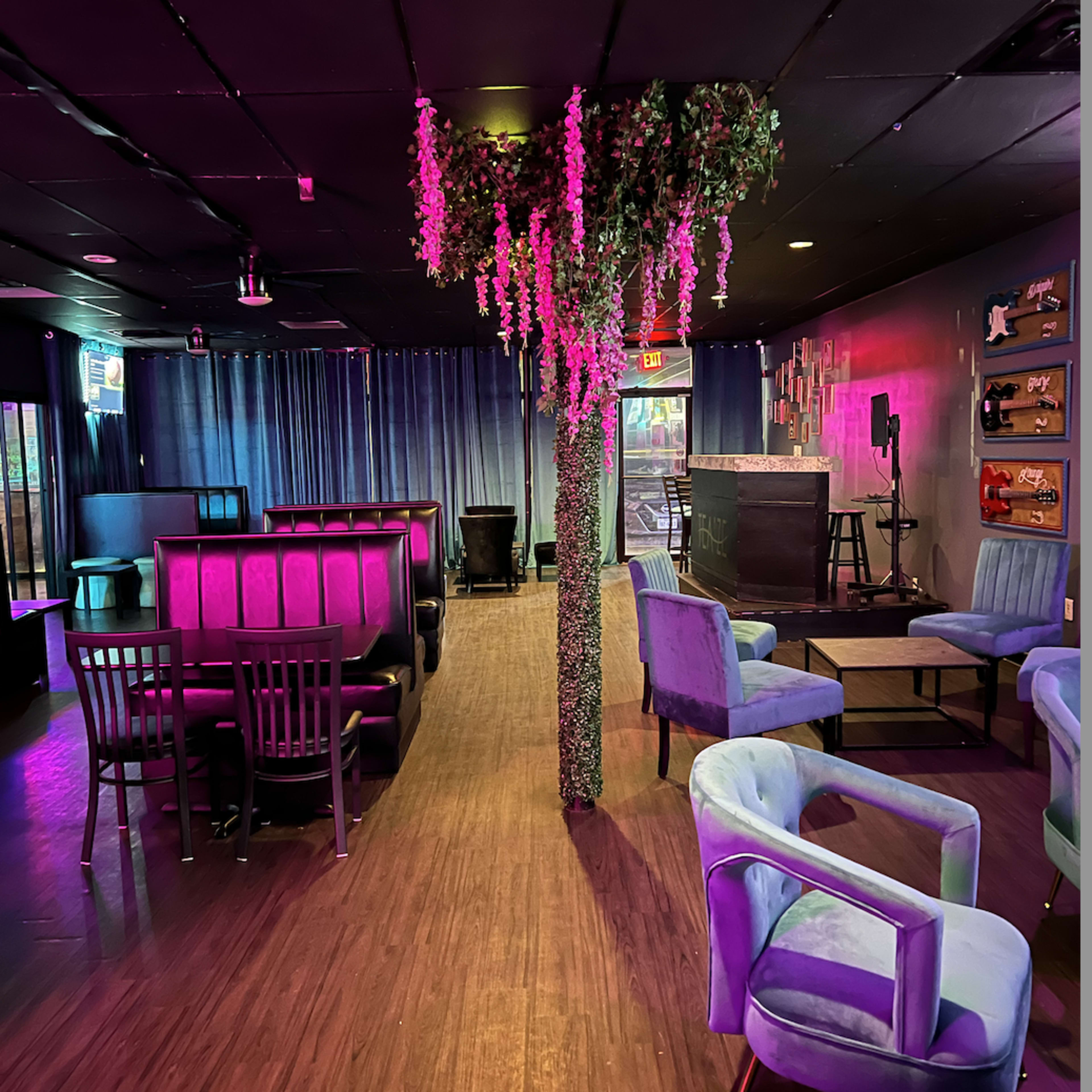 The interior of a lounge features a central plant column surrounded by seating areas with blue chairs and dark booths, illuminated by colorful lights.