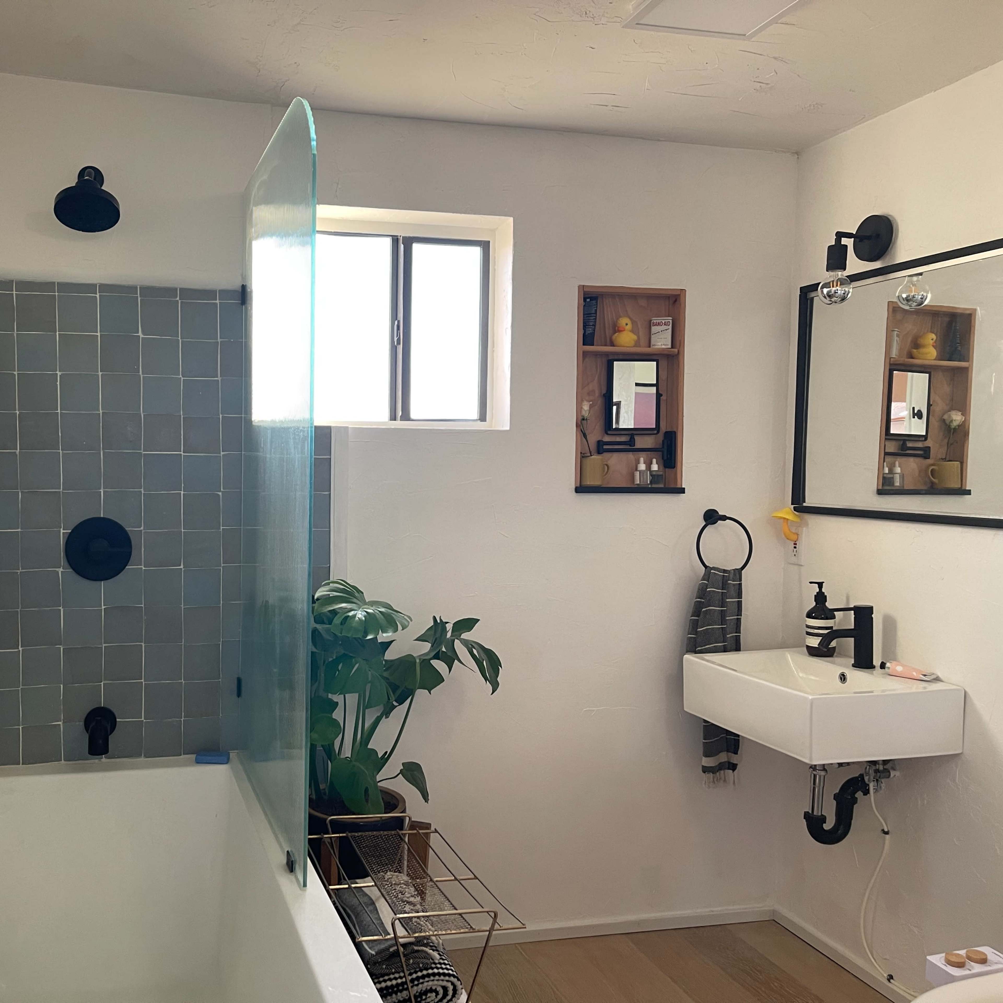 A modern bathroom with a bathtub, a wall-mounted sink, and a window, featuring tiled walls and greenery.