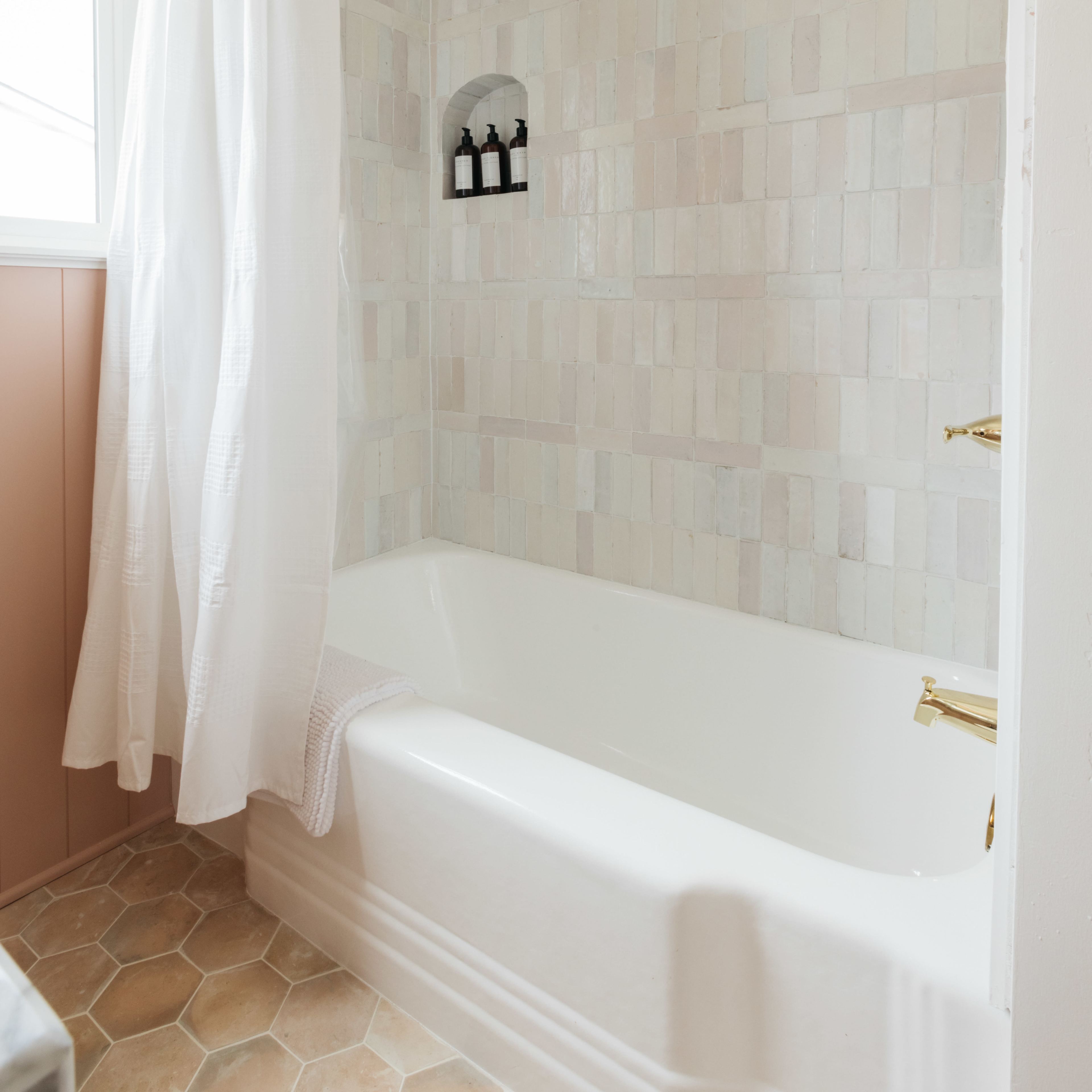A bathroom with a white bathtub, beige hexagonal tiled floor, and a shower area enclosed by a white curtain, with light-colored, patterned tiled walls.