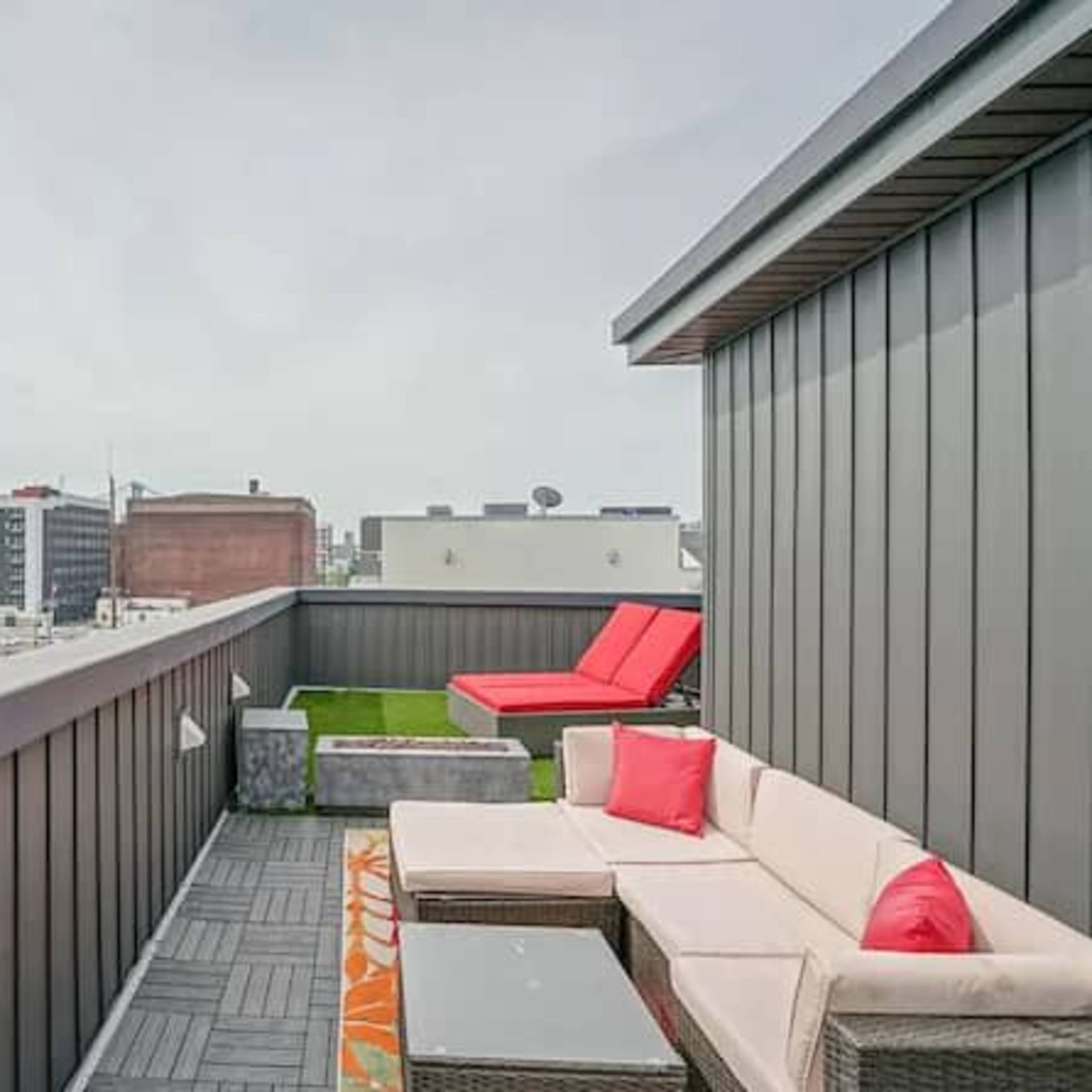A rooftop terrace features a sectional sofa, a red lounge chair, and a view of the city skyline and a bridge.