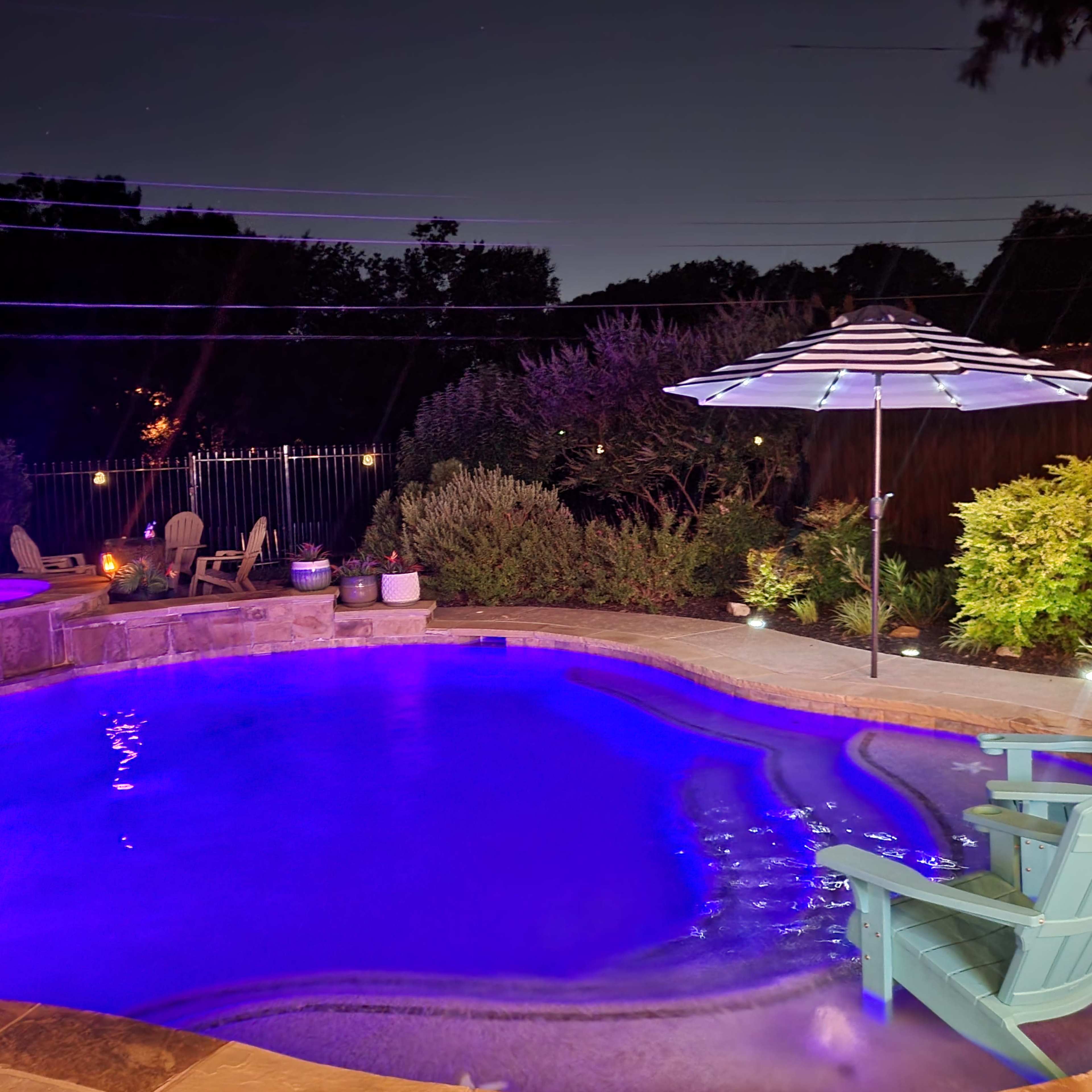 A illuminated swimming pool at night, featuring a lounge chair and an umbrella beside it, surrounded by landscaped greenery.