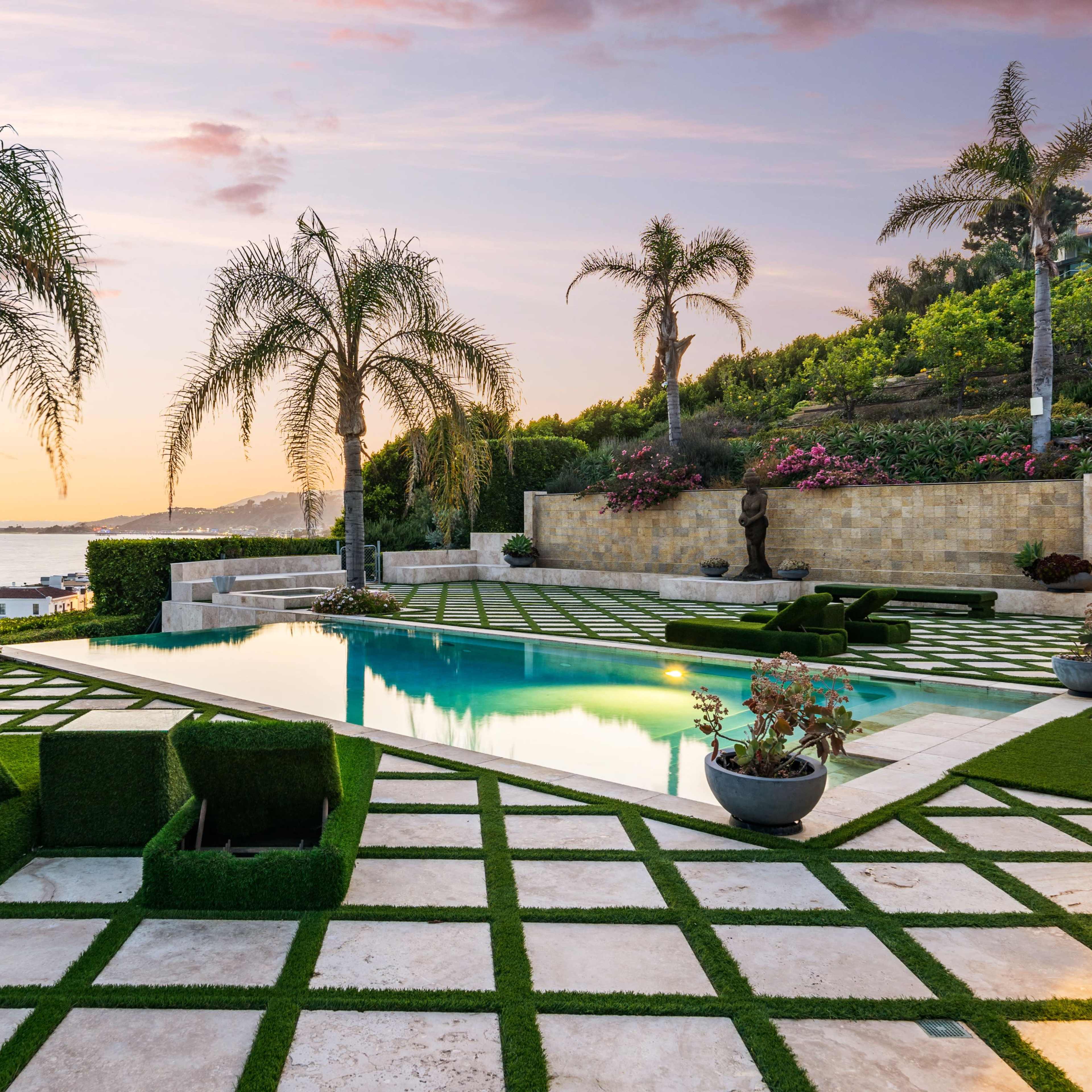 A landscaped outdoor area featuring a swimming pool surrounded by palm trees and a checkerboard pattern of grass and stone tiles at sunset.