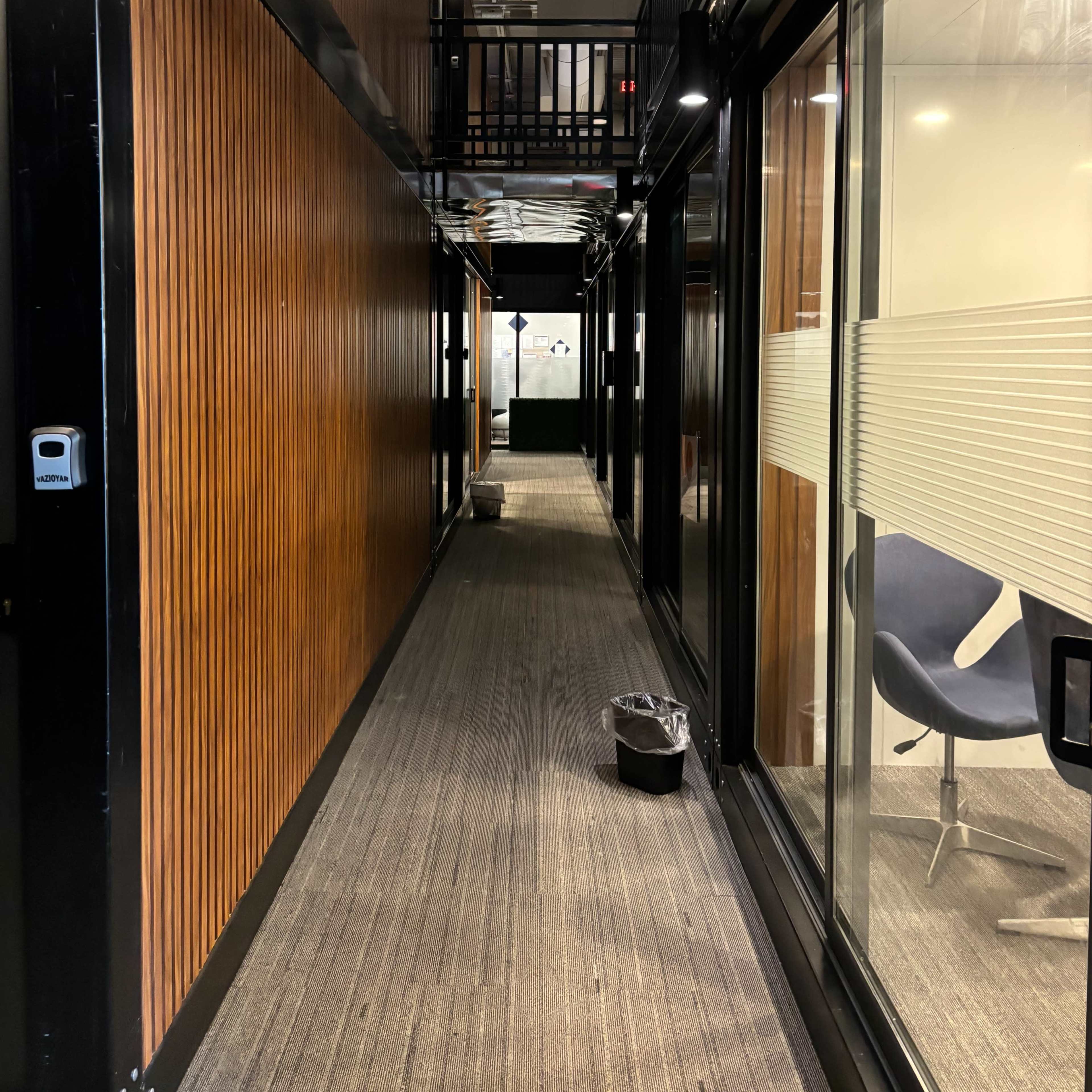 A narrow hallway lined with wood-paneled walls, leading to a glass-walled meeting area and featuring two trash bins along the floor.