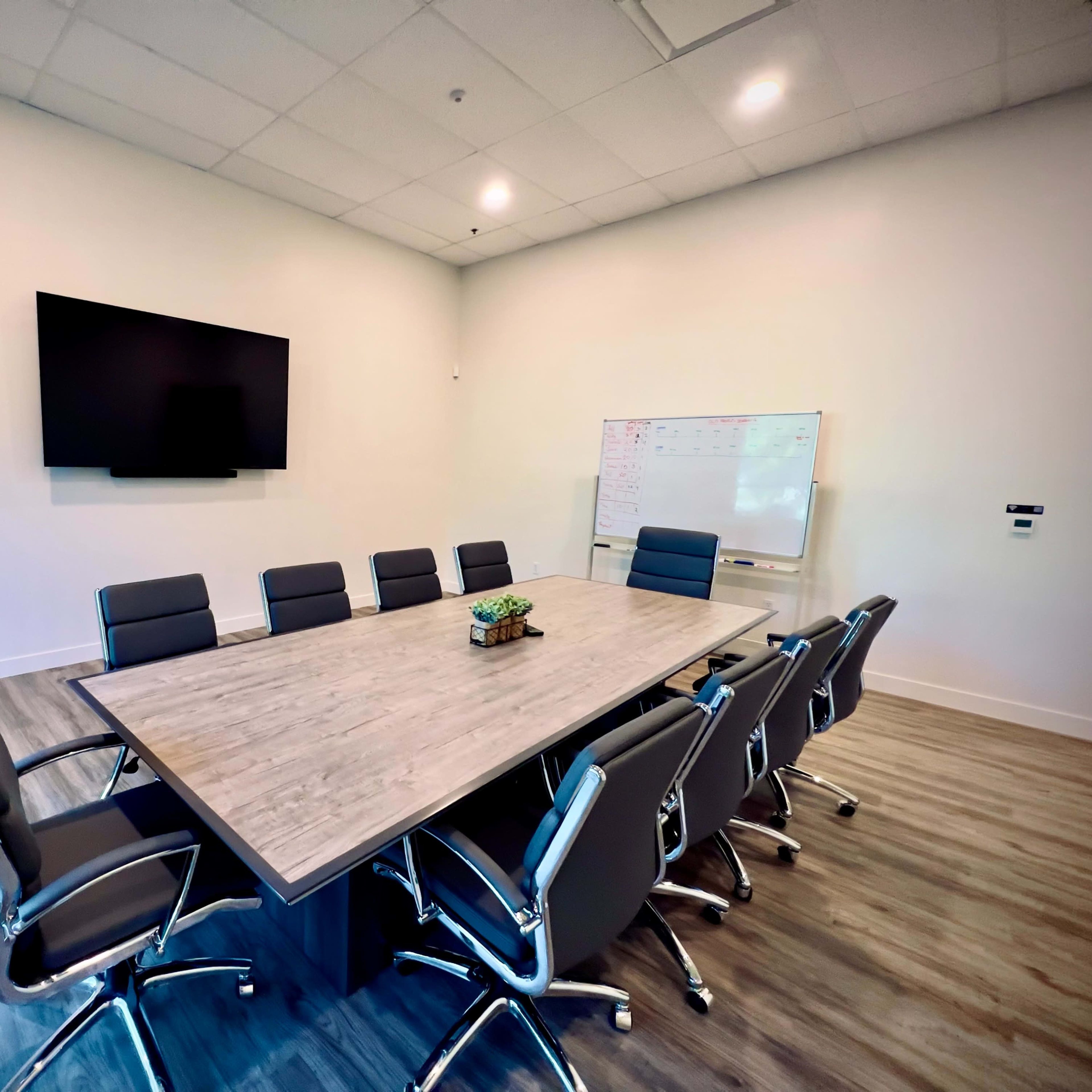 A modern conference room features a large wooden table surrounded by sleek, black chairs, with a wall-mounted screen and a whiteboard nearby.