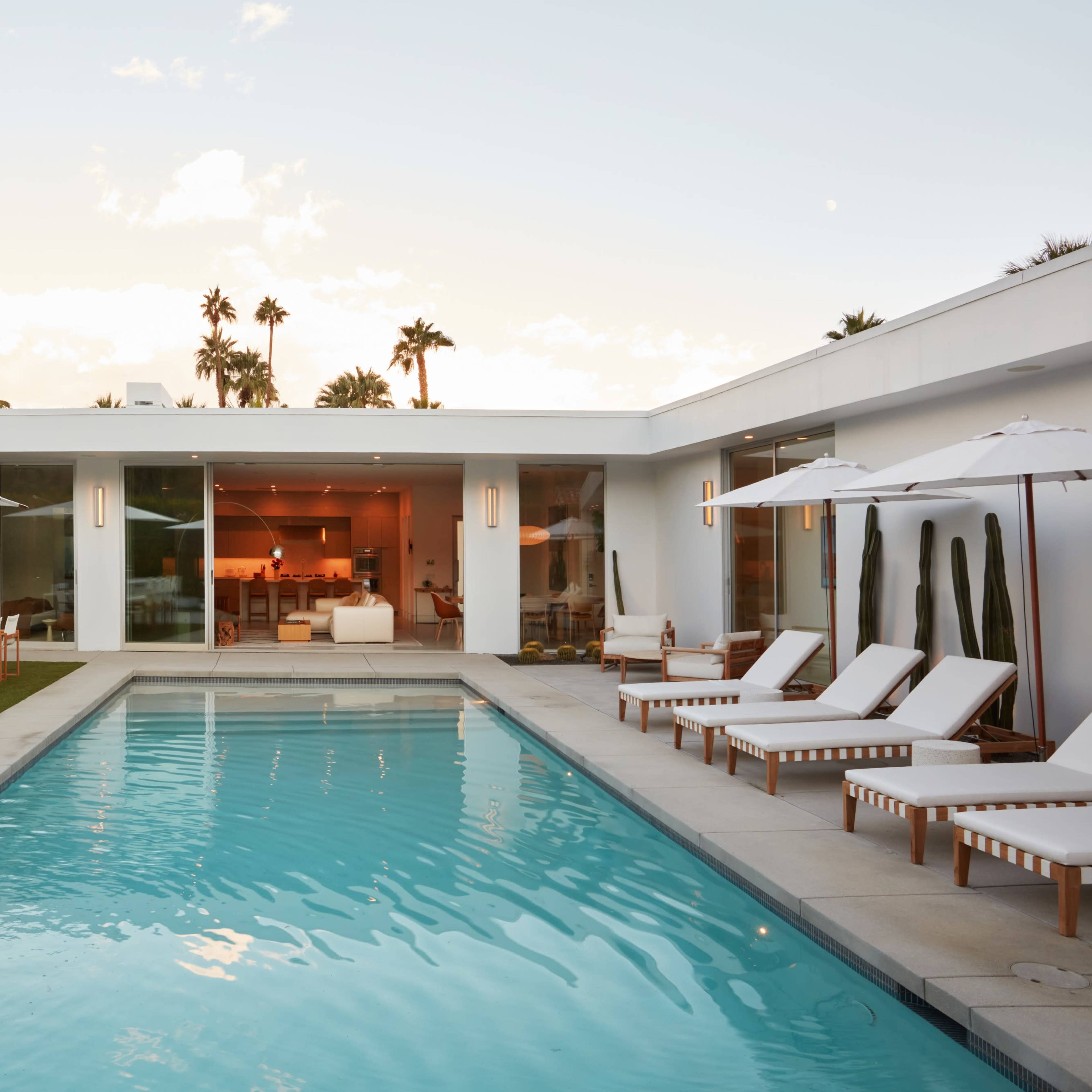 A modern home with large glass walls surrounds a rectangular swimming pool, featuring lounge chairs and patio seating under white umbrellas, set against a backdrop of palm trees.