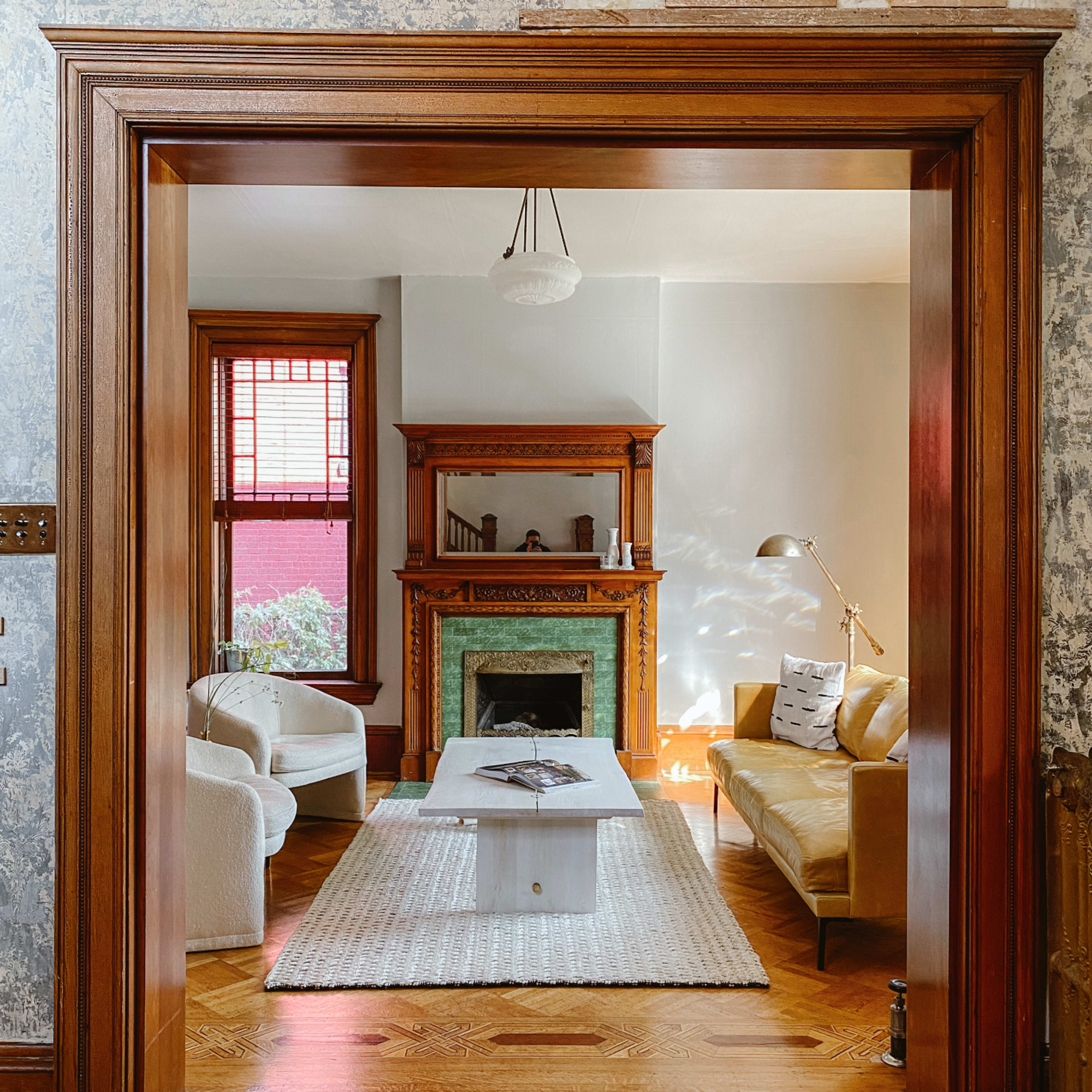 A cozy living room features a yellow sofa, white armchairs, a decorative fireplace, and a coffee table on a woven rug, framed by a wooden archway.