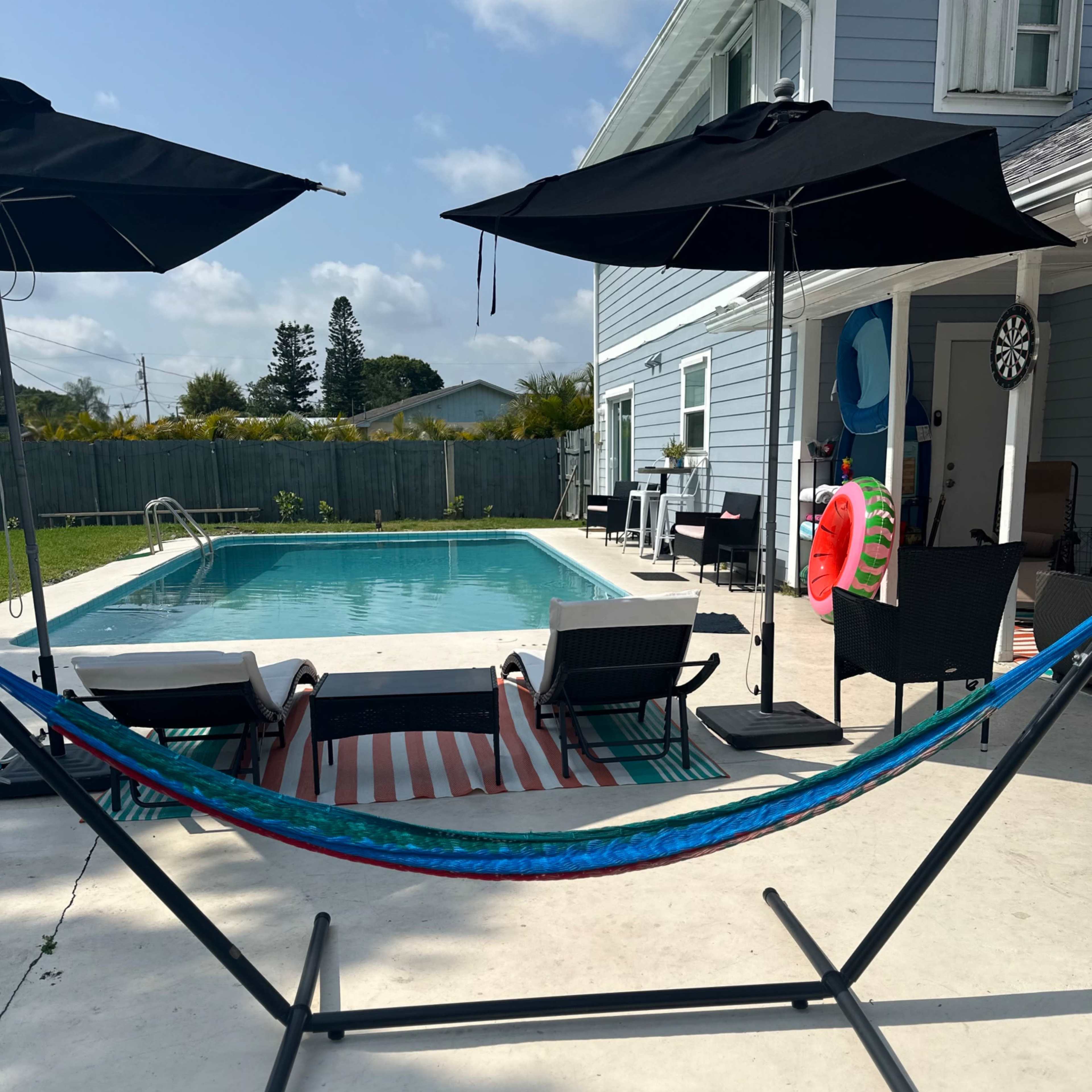 A hammock is set up near a swimming pool, surrounded by lounge chairs and patio umbrellas at a residential property.