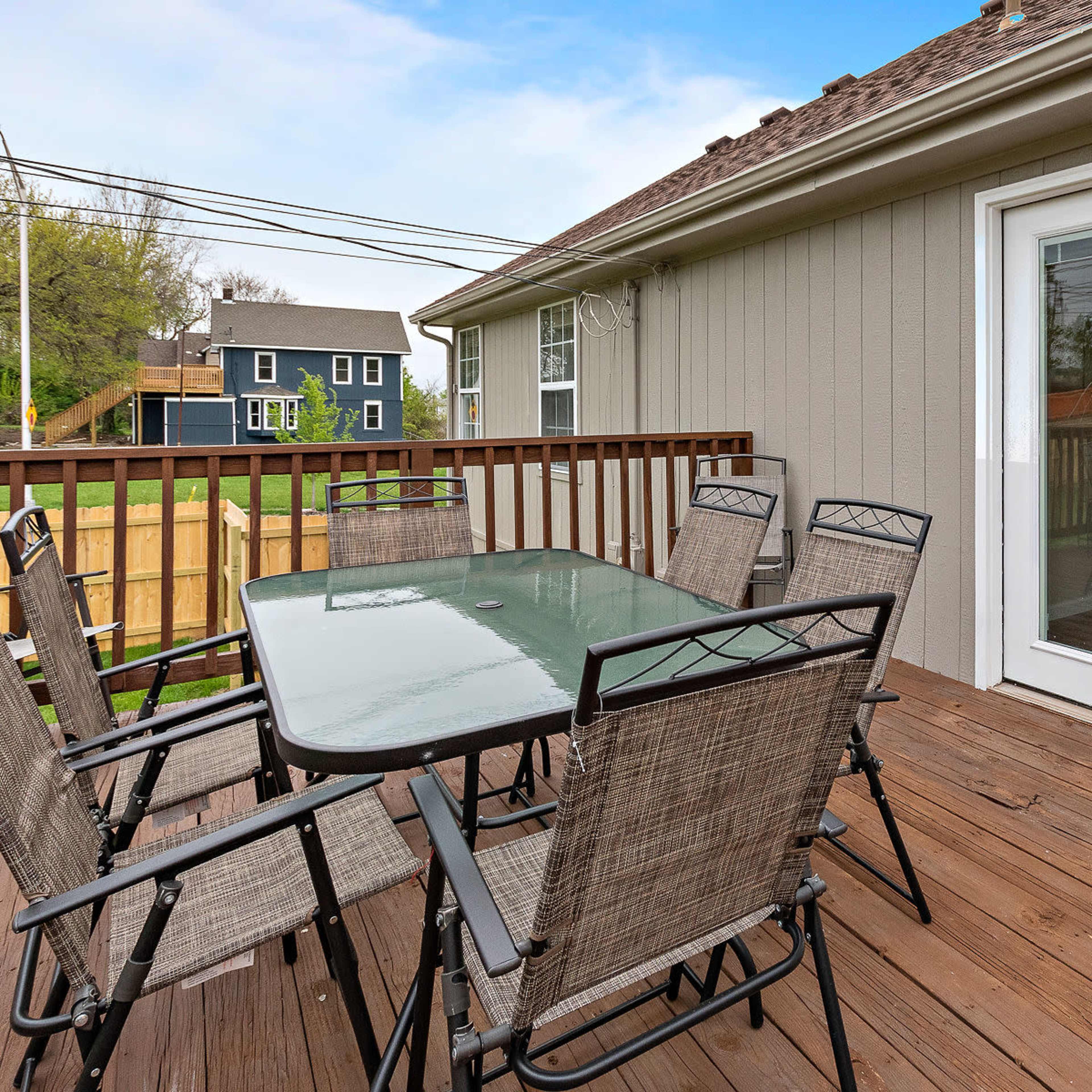 A wooden patio features a glass-top dining table surrounded by six chairs, leading to double doors that open into a house.