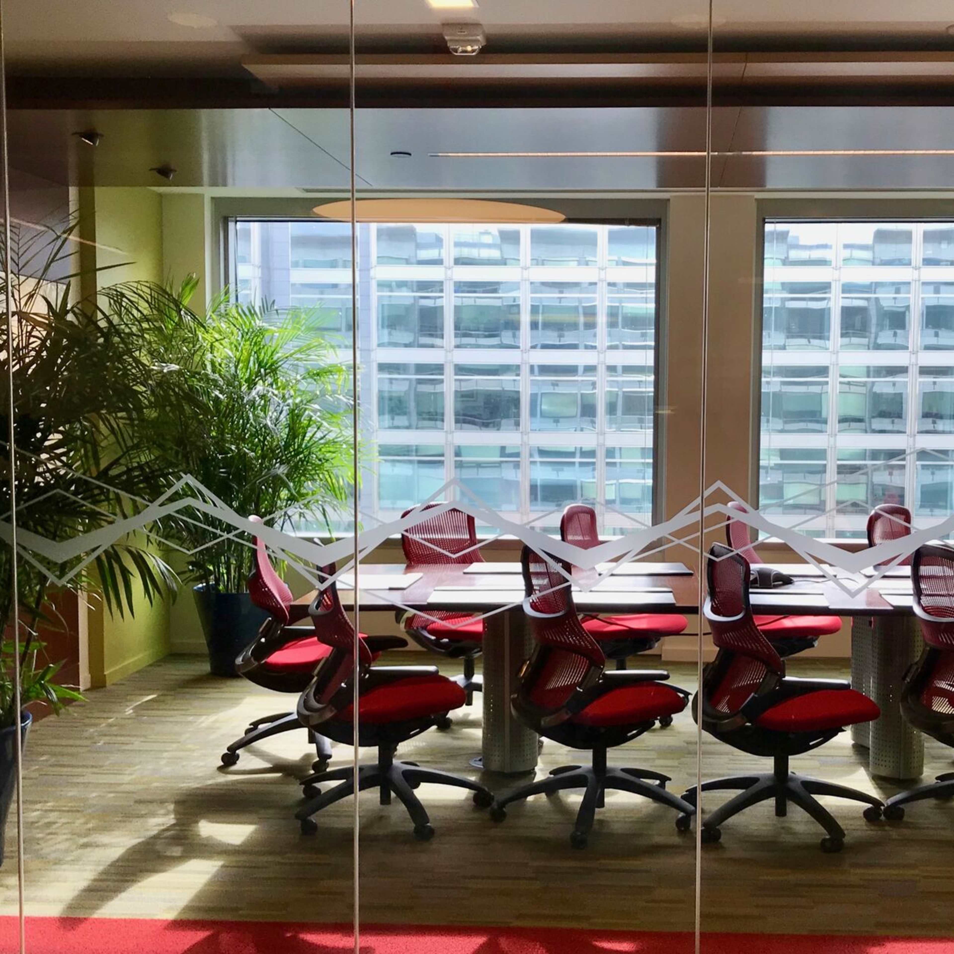 A conference room with a long table surrounded by red office chairs and large windows overlooking a modern building.