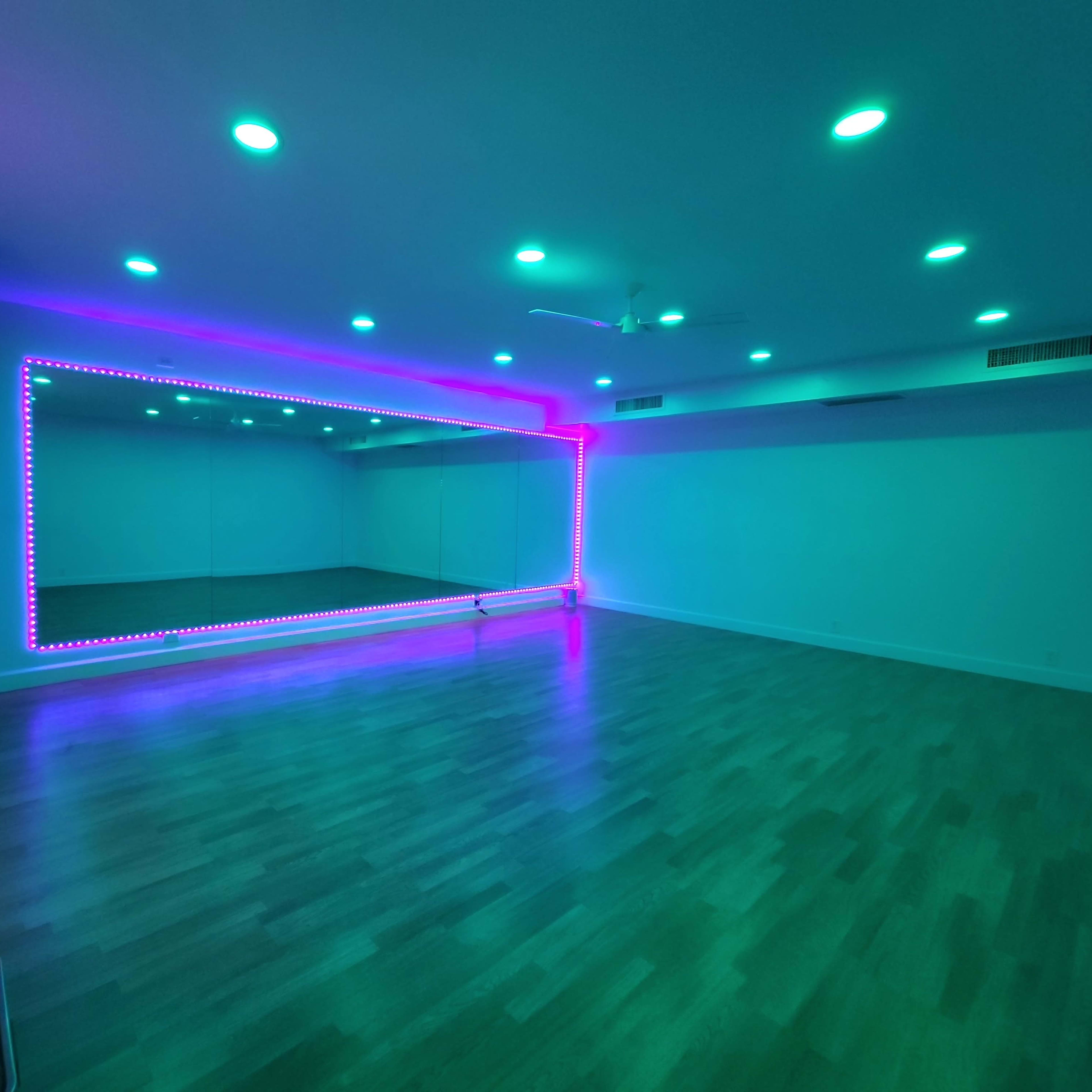 A spacious room with polished wood flooring and a mirrored wall framed by colorful LED lights.
