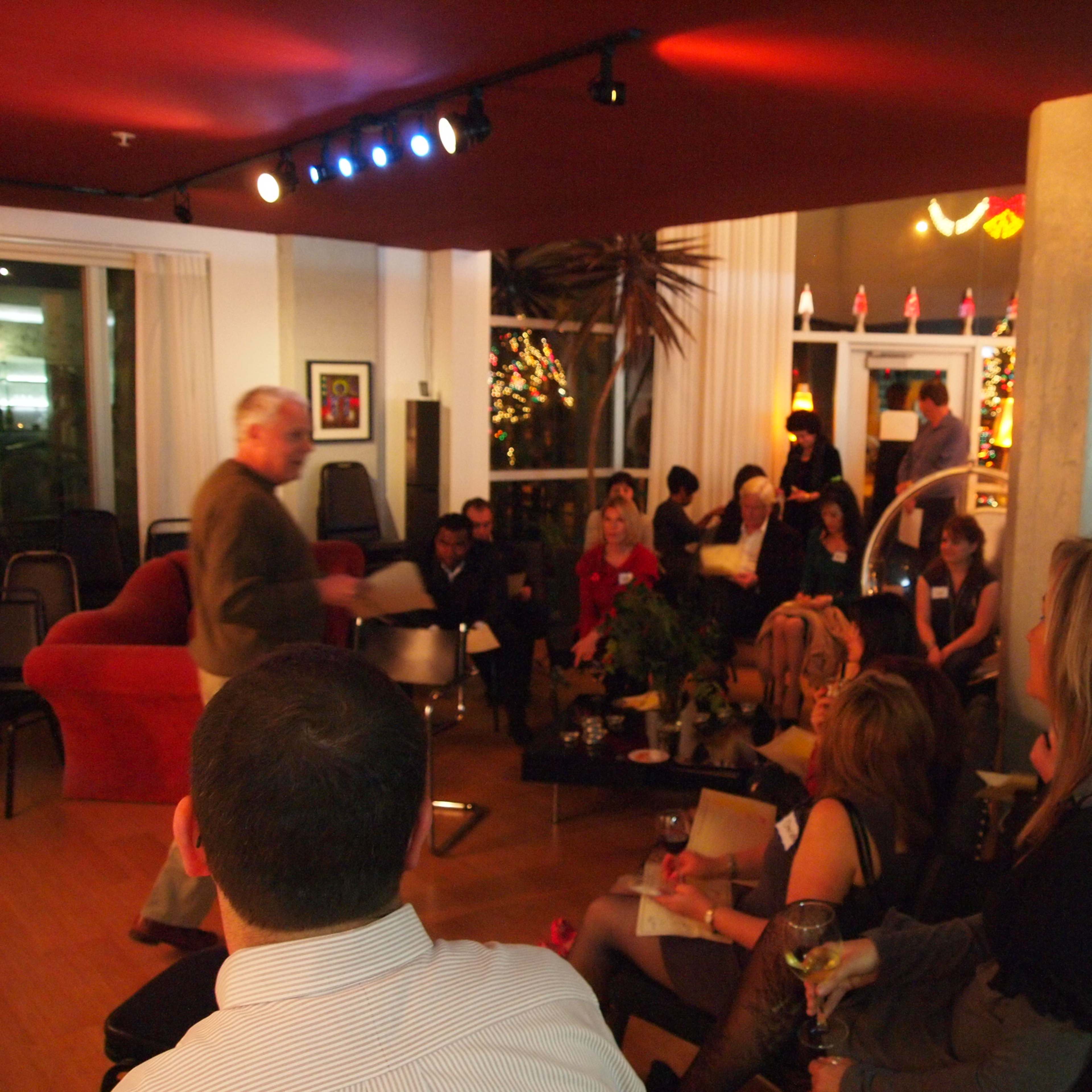 A group of people is seated in a well-lit indoor space, with a speaker presenting at the front and decorations including plants and lights in the background.