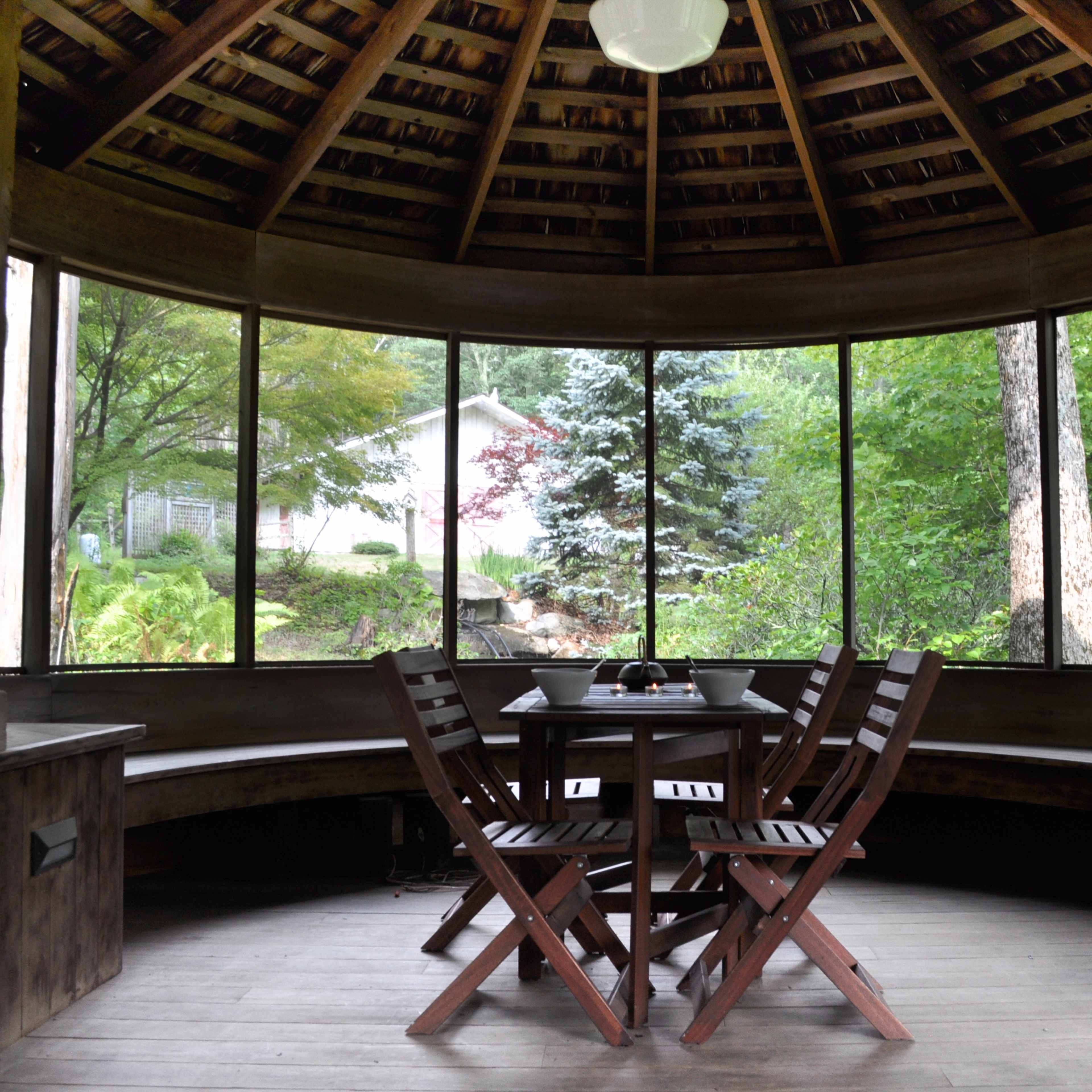 A circular wooden gazebo with large windows, featuring a table surrounded by four wooden chairs.