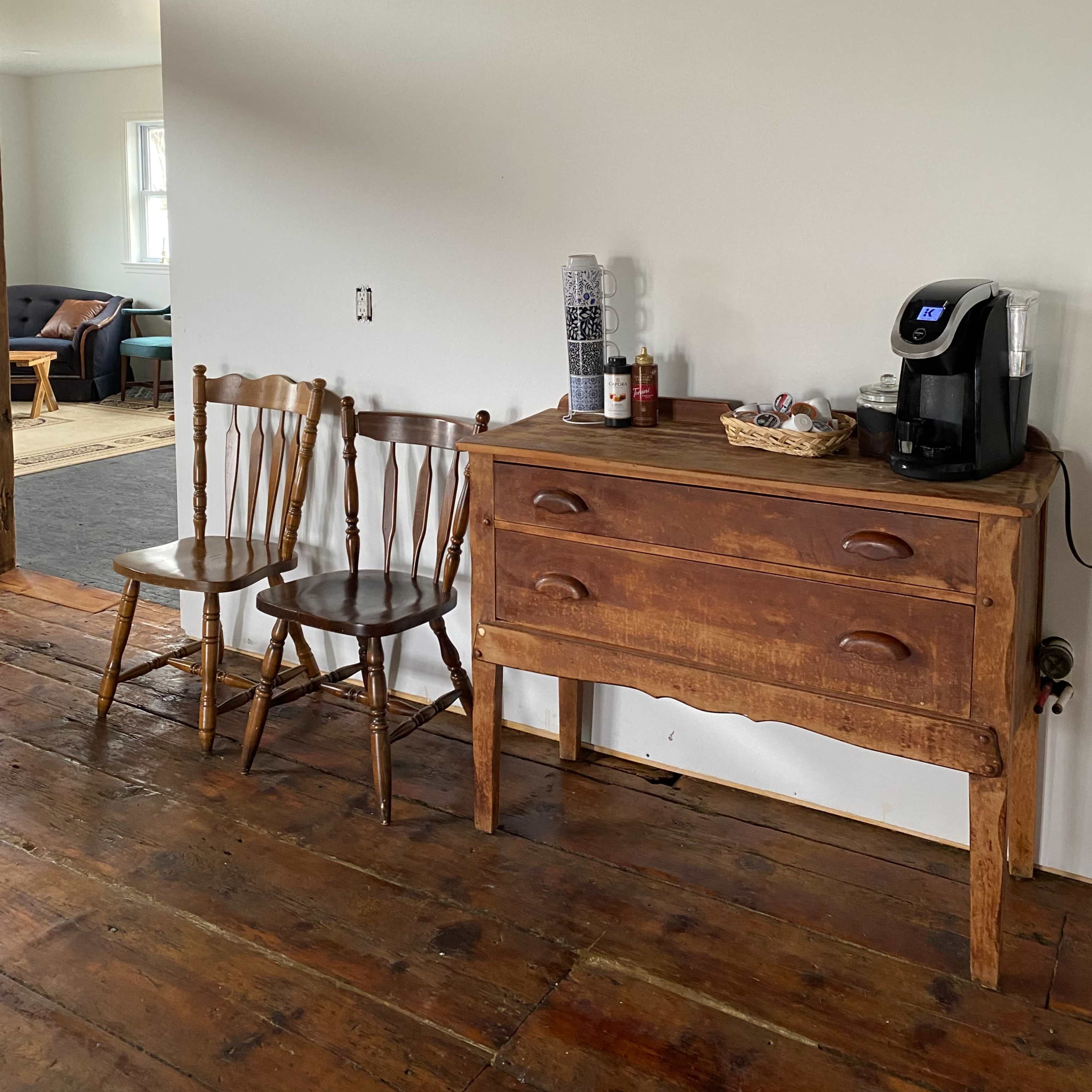 A wooden side table with two chairs and a coffee maker is situated against a white wall in a room with wooden flooring.