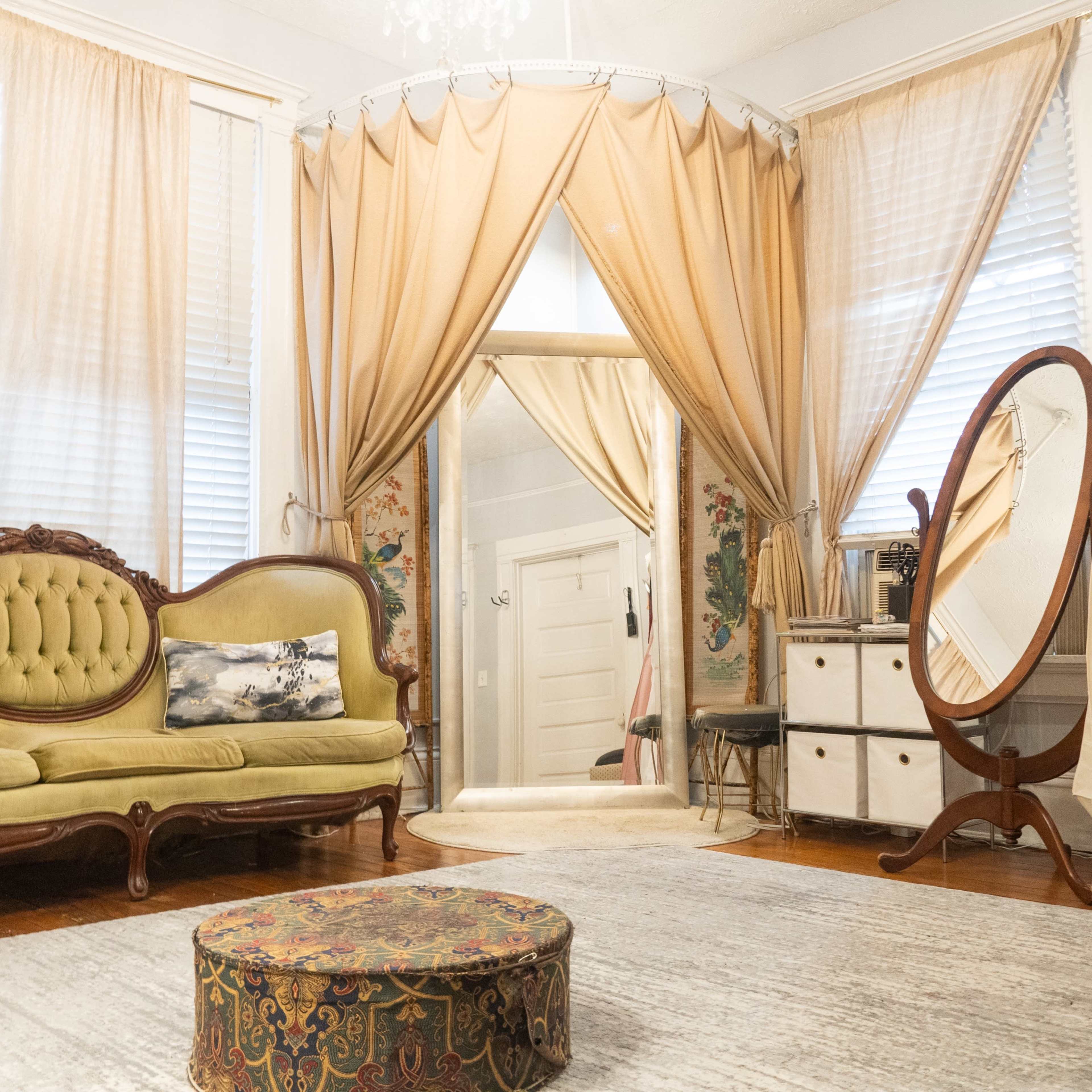 A well-furnished room featuring a green sofa, a round decorative ottoman, a large mirror, and draped curtains.