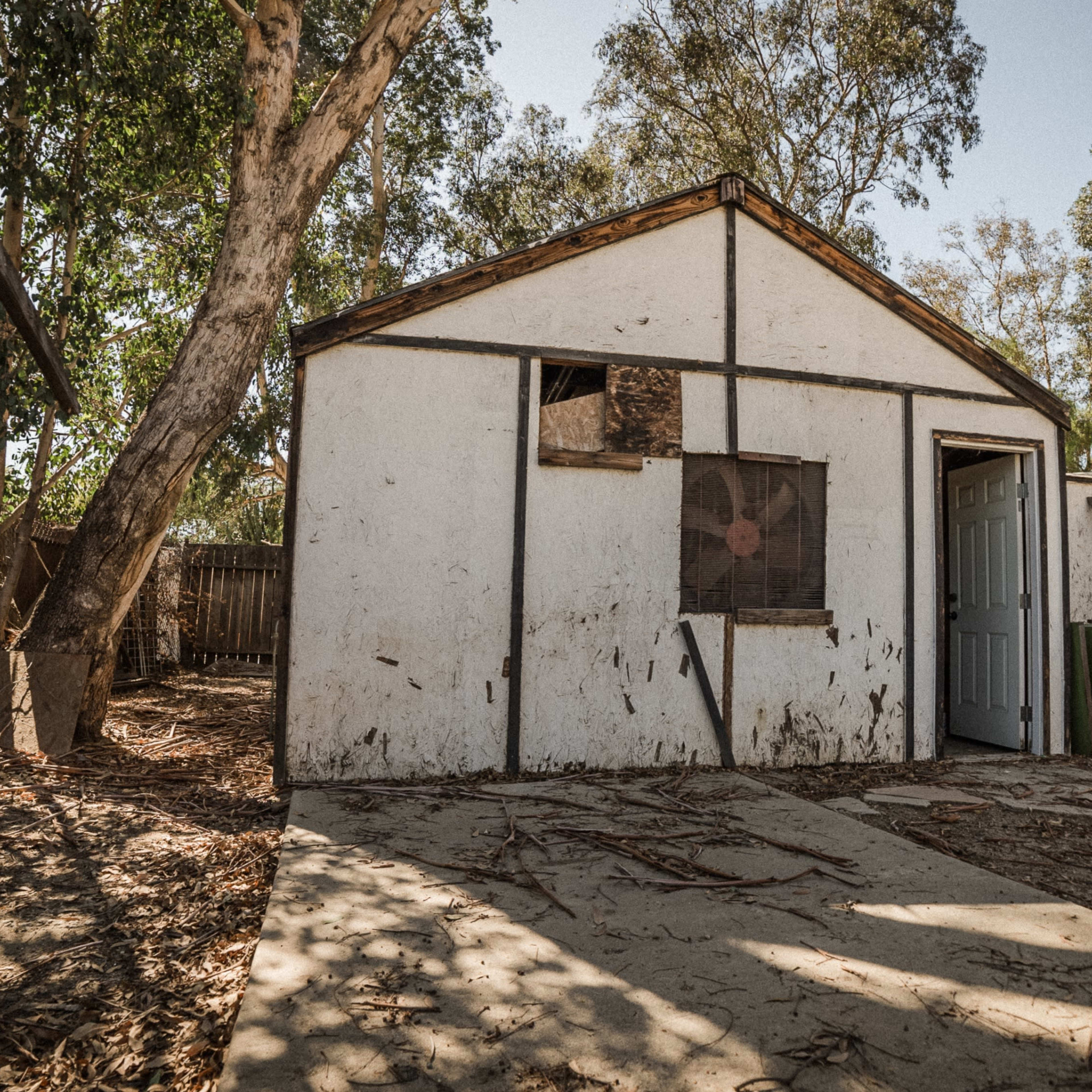 A weathered, white shed with a door and a window, surrounded by dirt and sparse vegetation.