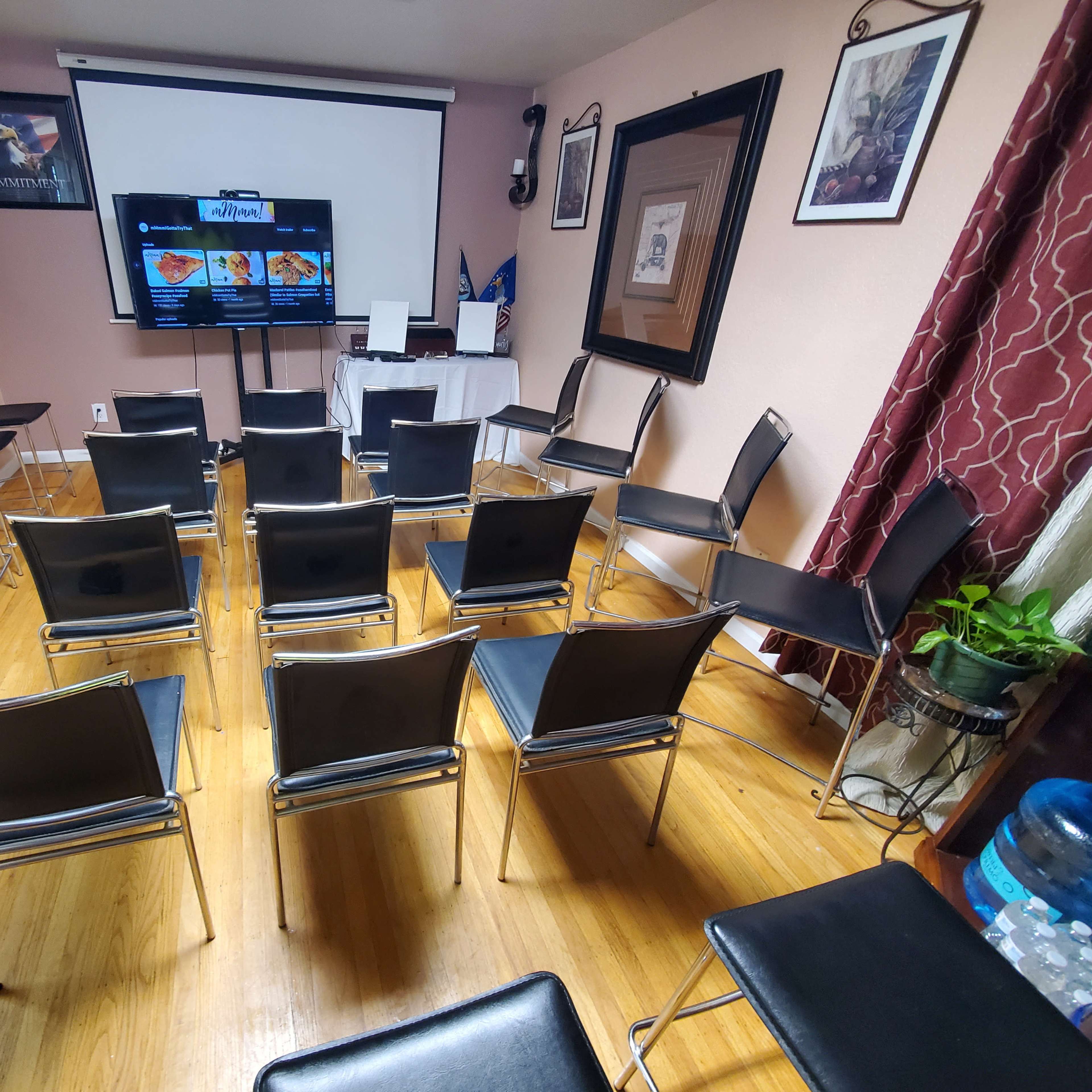 A room is set up with rows of black chairs facing a screen displaying content, alongside a whiteboard and decorative elements on the walls.