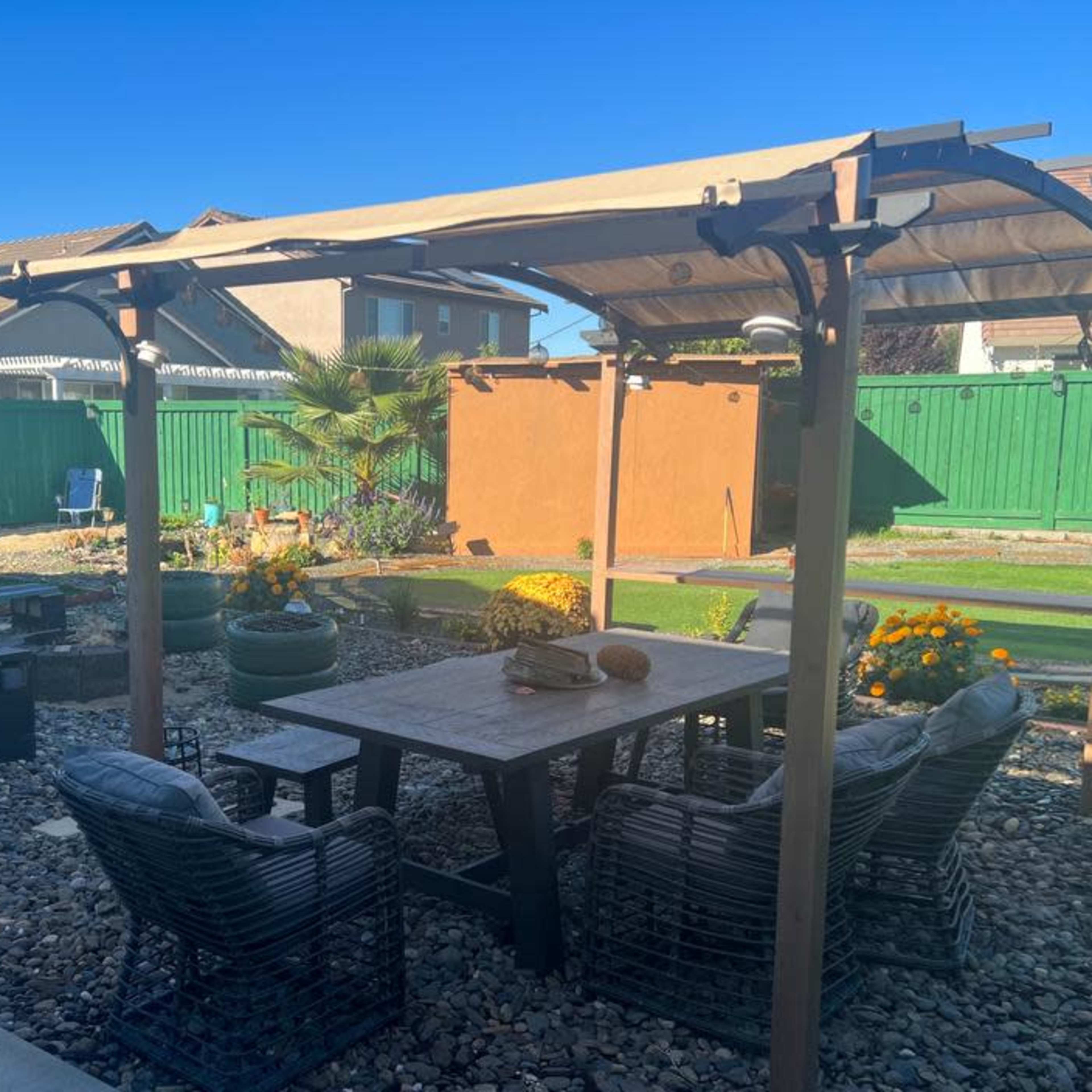 A shaded patio area features a table and chairs surrounded by decorative stones and plants in a residential backyard.