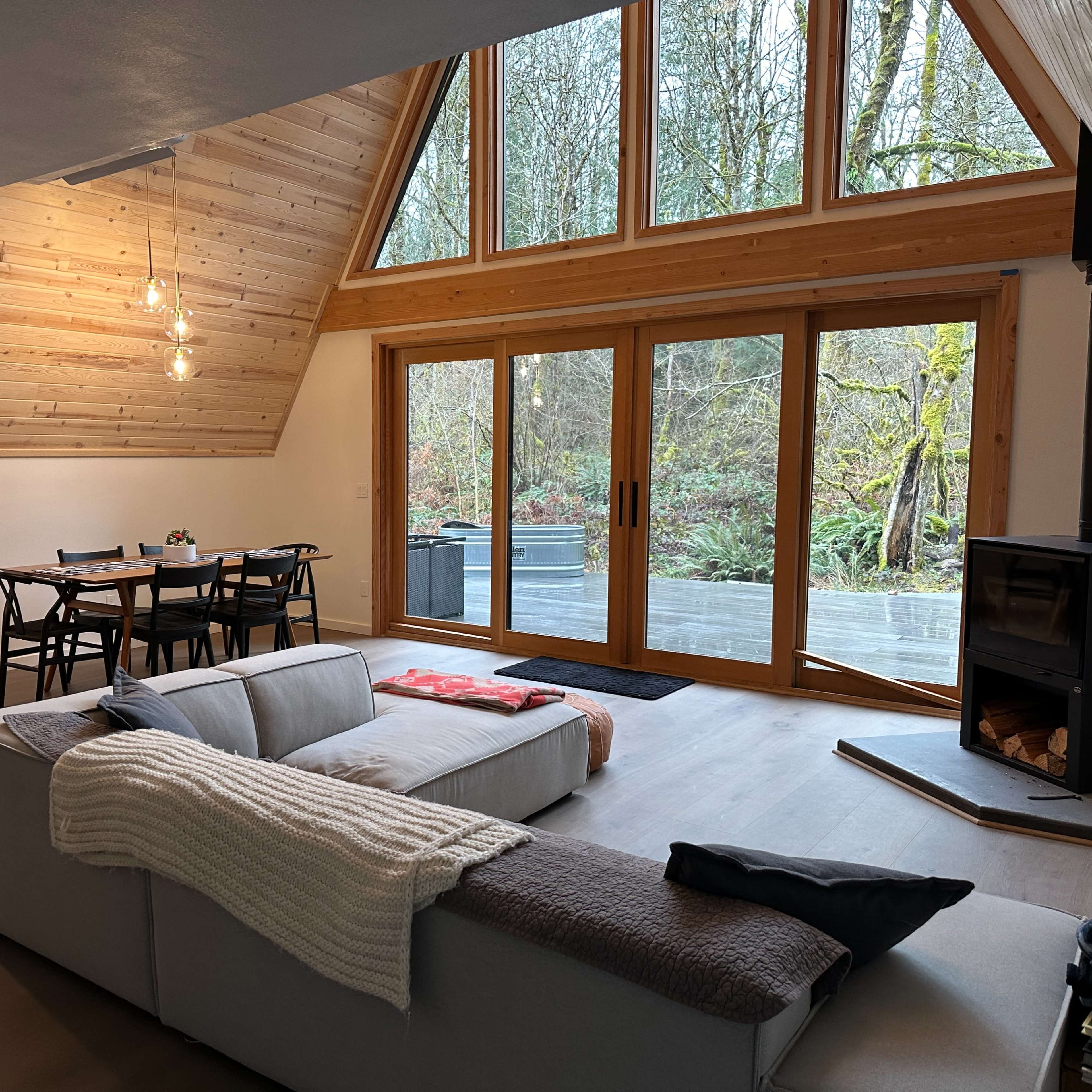 A modern living space featuring a large window with wooden trim, a cozy sofa, a dining table, and a wood-burning stove, all within a cabin-like interior design.