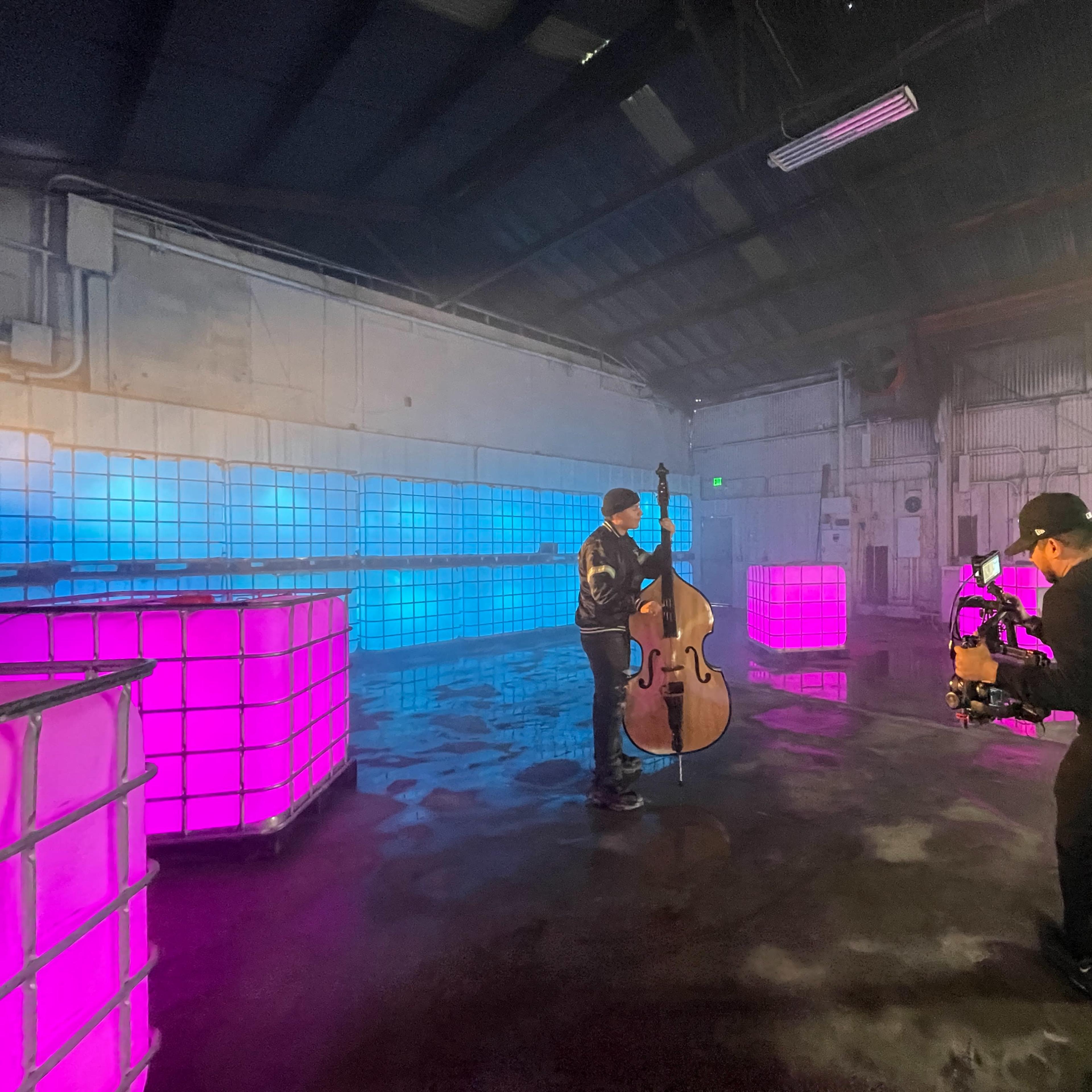A musician plays a double bass in a foggy, industrial space with brightly lit cube structures.