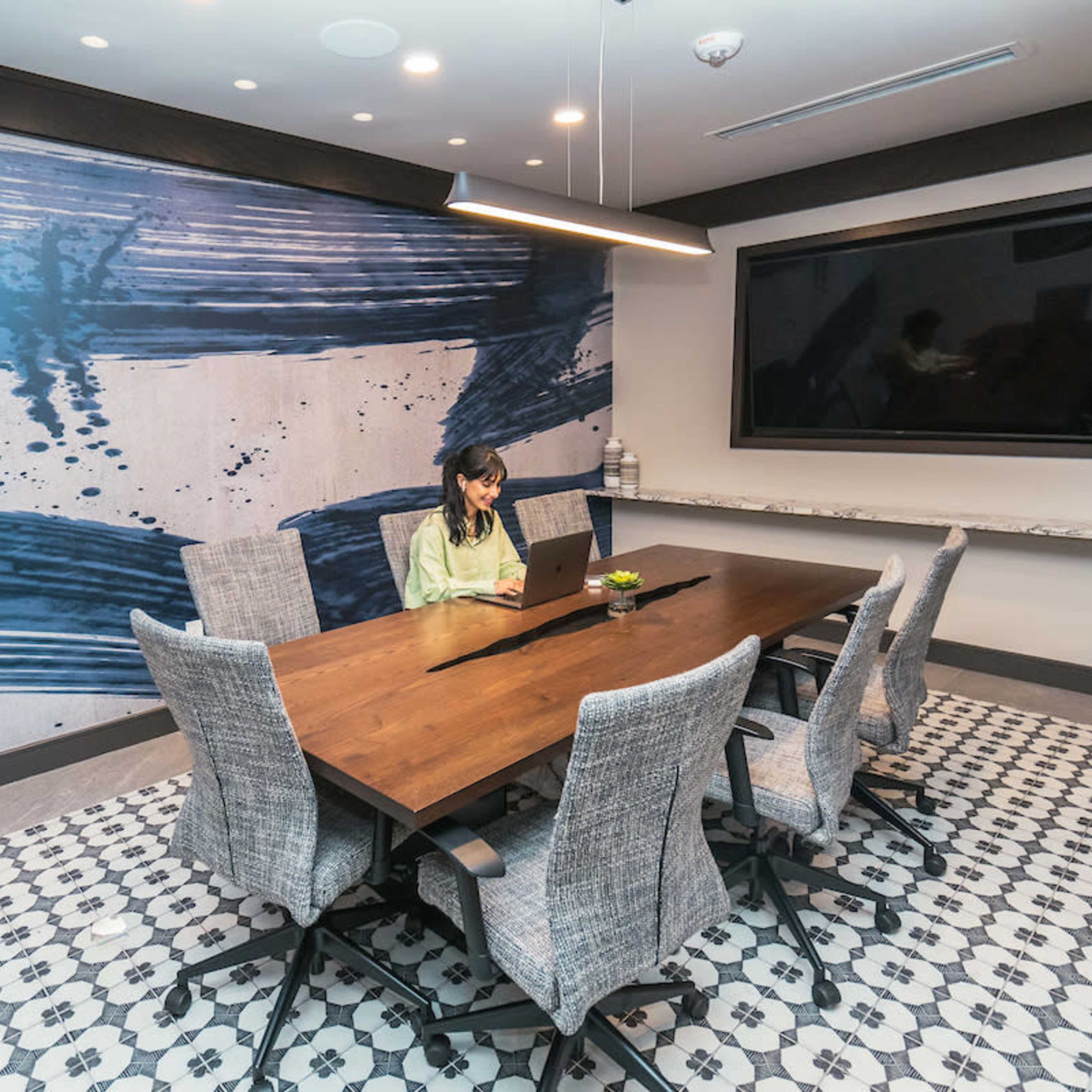 A woman is working on a laptop at a wooden table in a meeting room with a blue abstract mural on the wall.