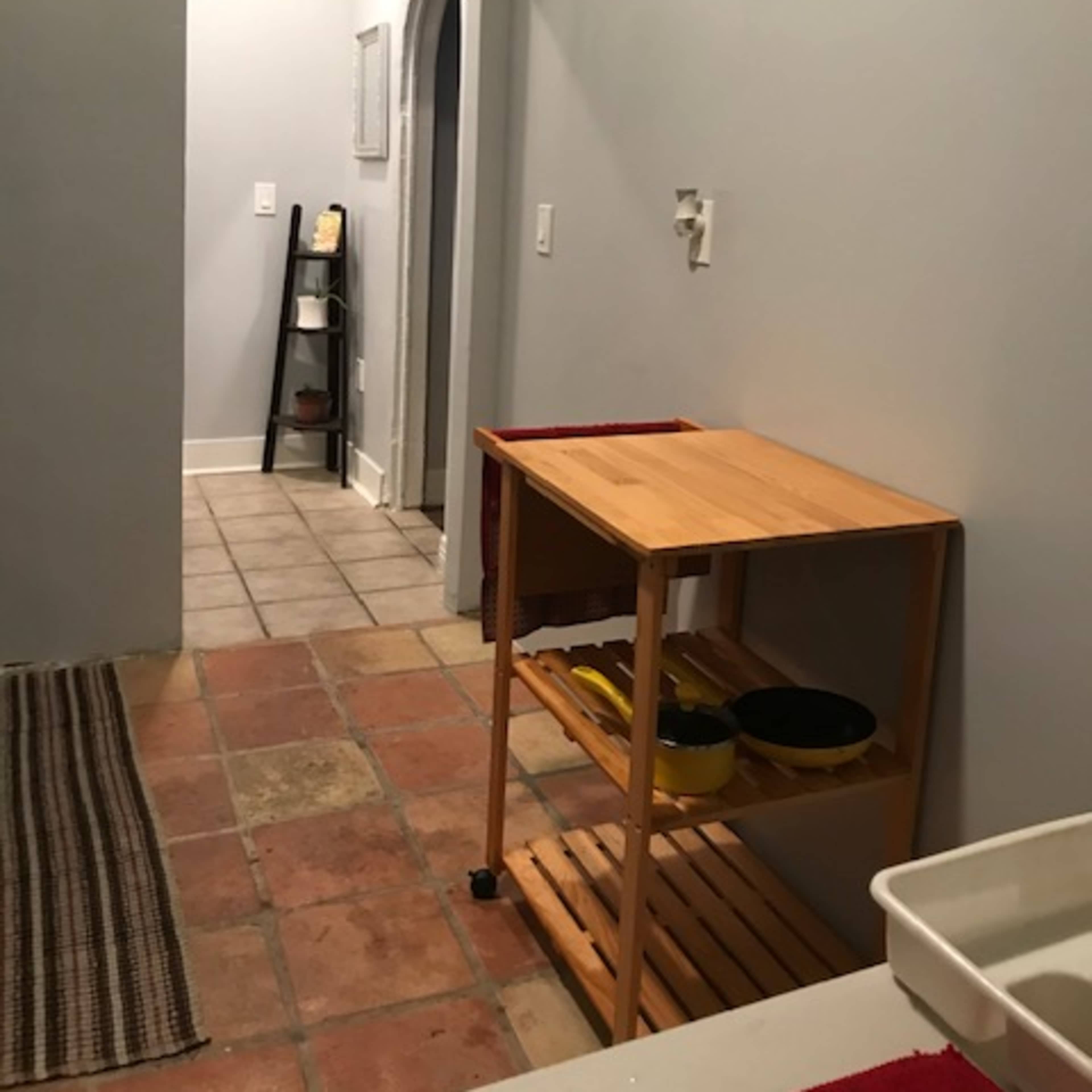 A narrow kitchen space with a wooden cart on the left, a tiled floor, and a doorway leading to a hallway in the background.