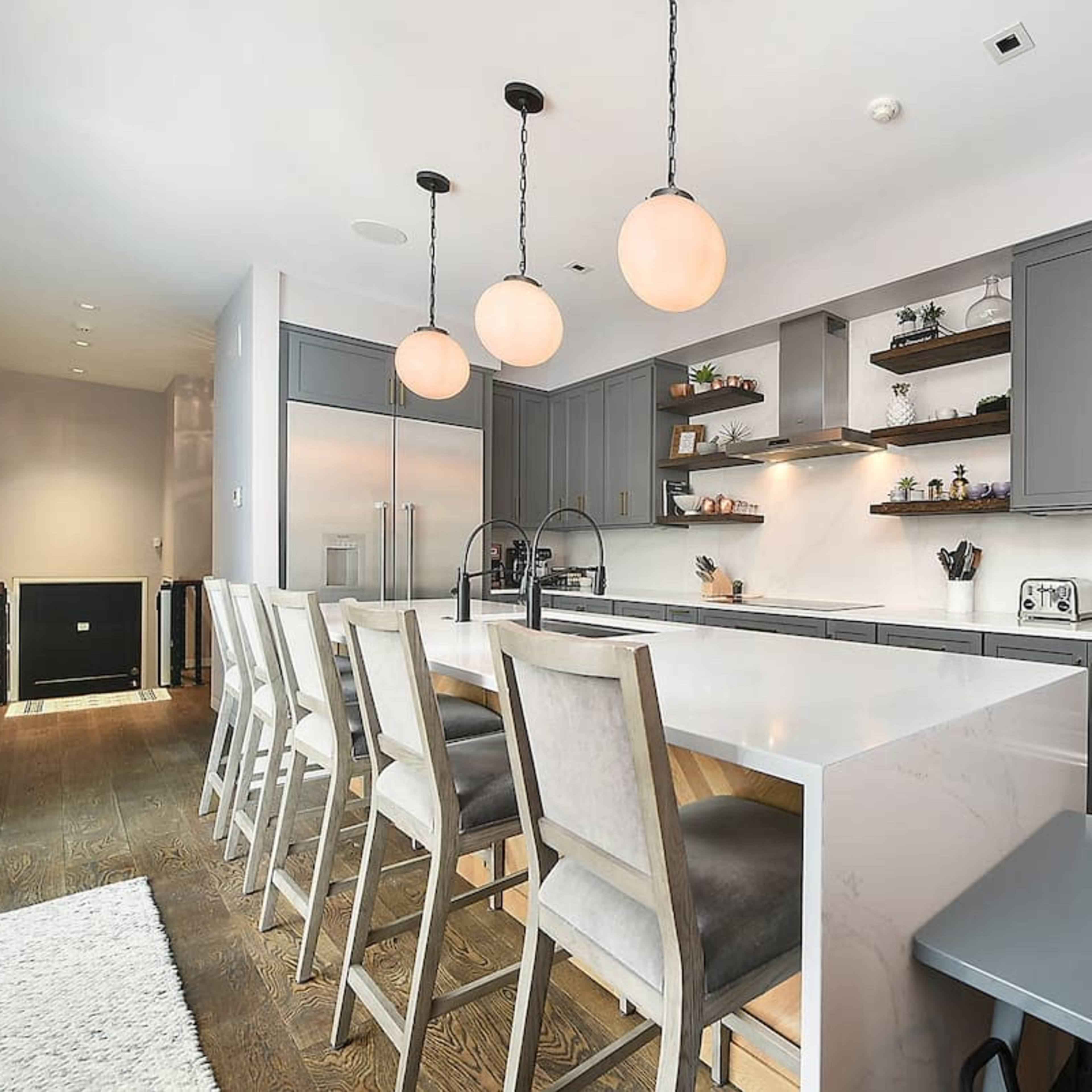 A modern kitchen features gray cabinetry, a large white island with bar seating, and pendant lights hung above.
