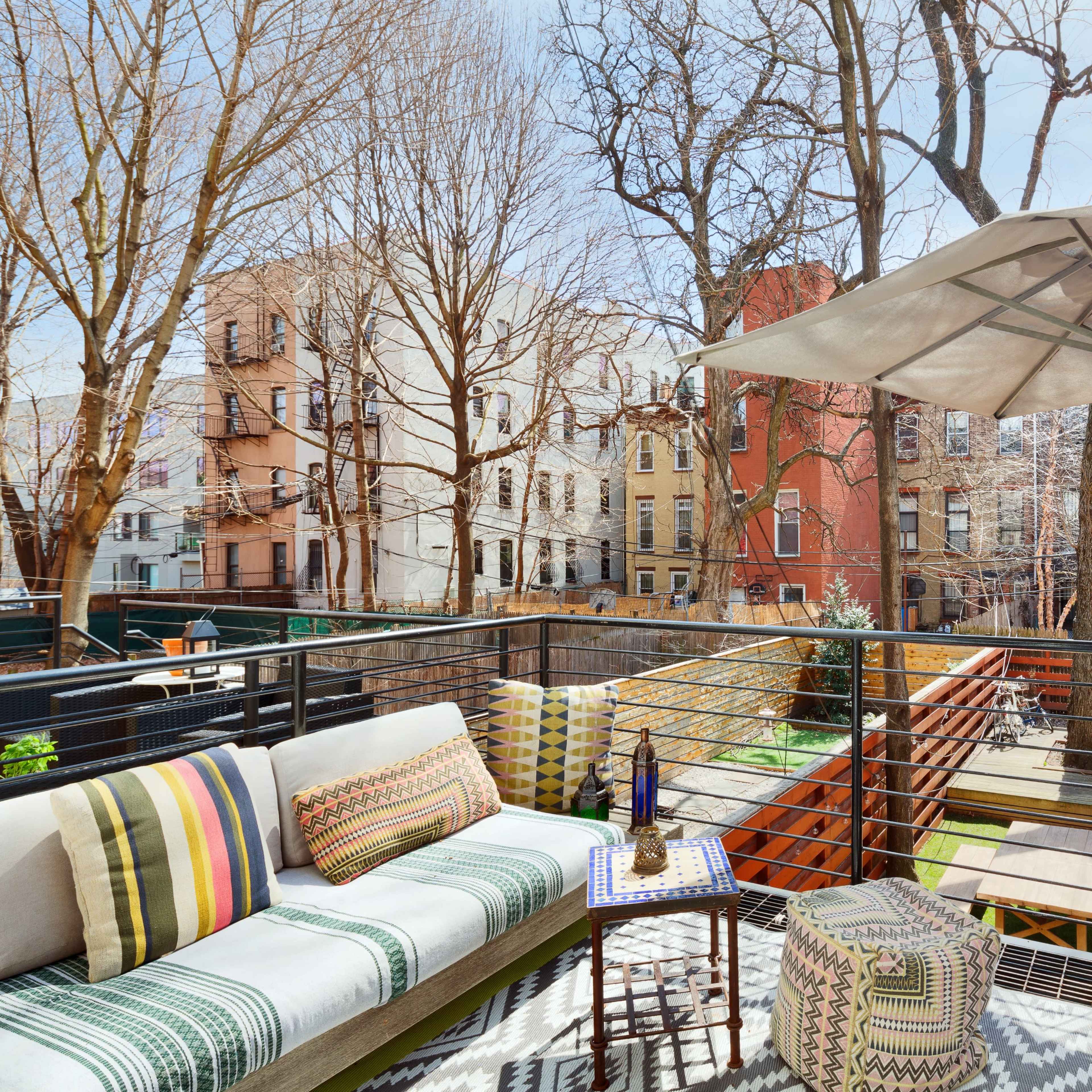 A terrace features a seating area with a sofa, umbrella, and small table overlooking a backyard with trees and buildings in the background.