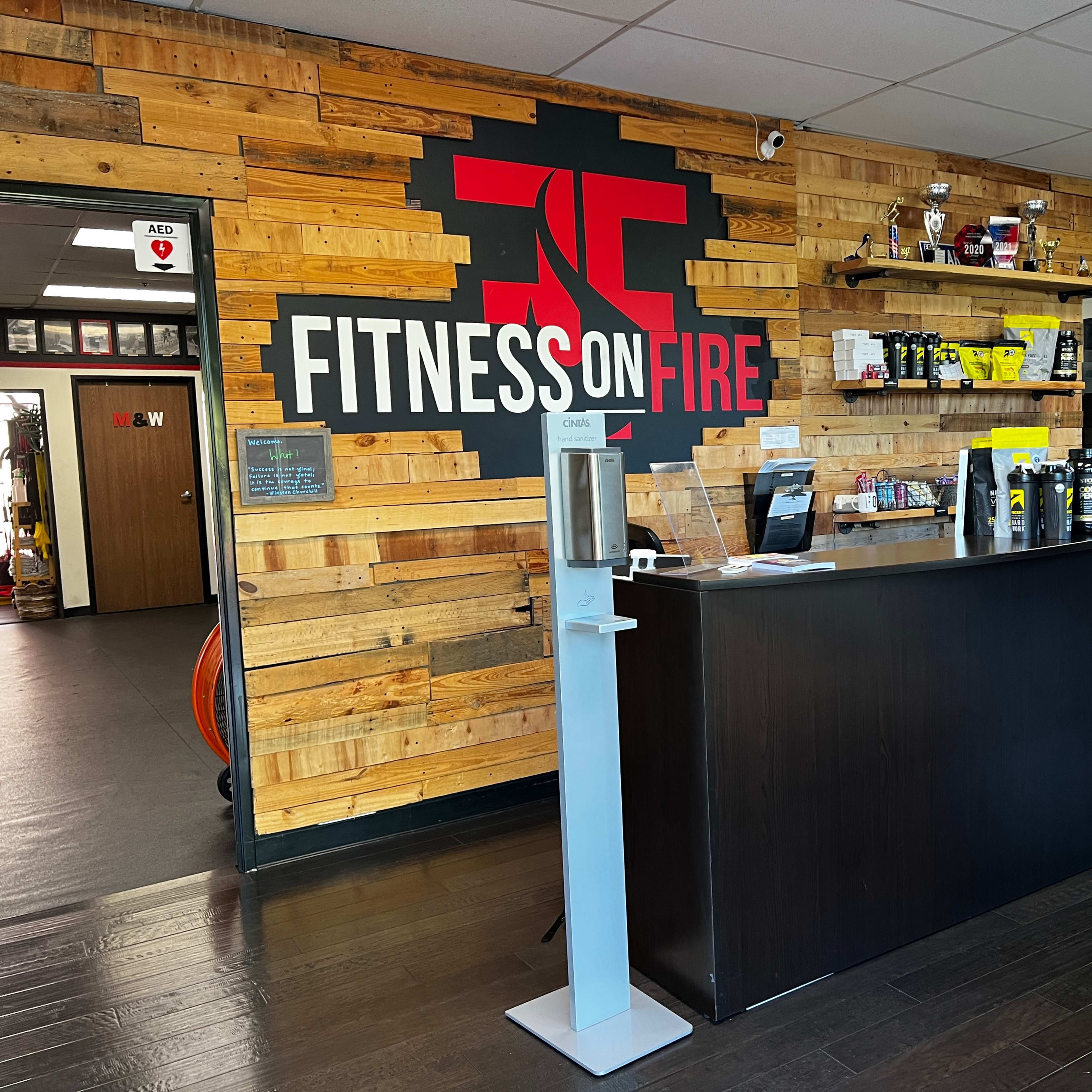 The interior of a fitness center features a reception desk with a backdrop of wooden planks and a large wall graphic that reads "FITNESS ON FIRE."