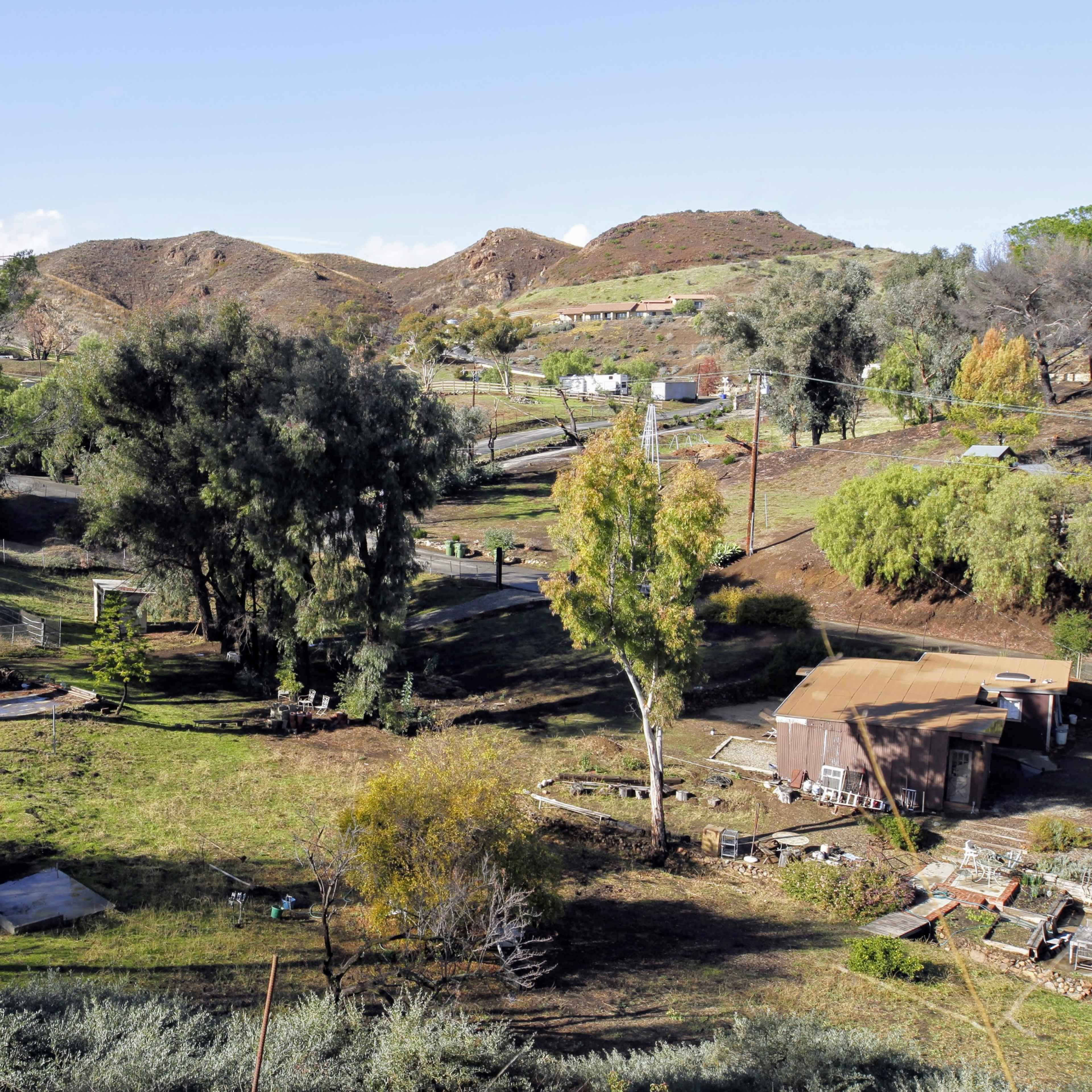A rural landscape with hills in the background, scattered trees, and a small homestead containing various structures and equipment.