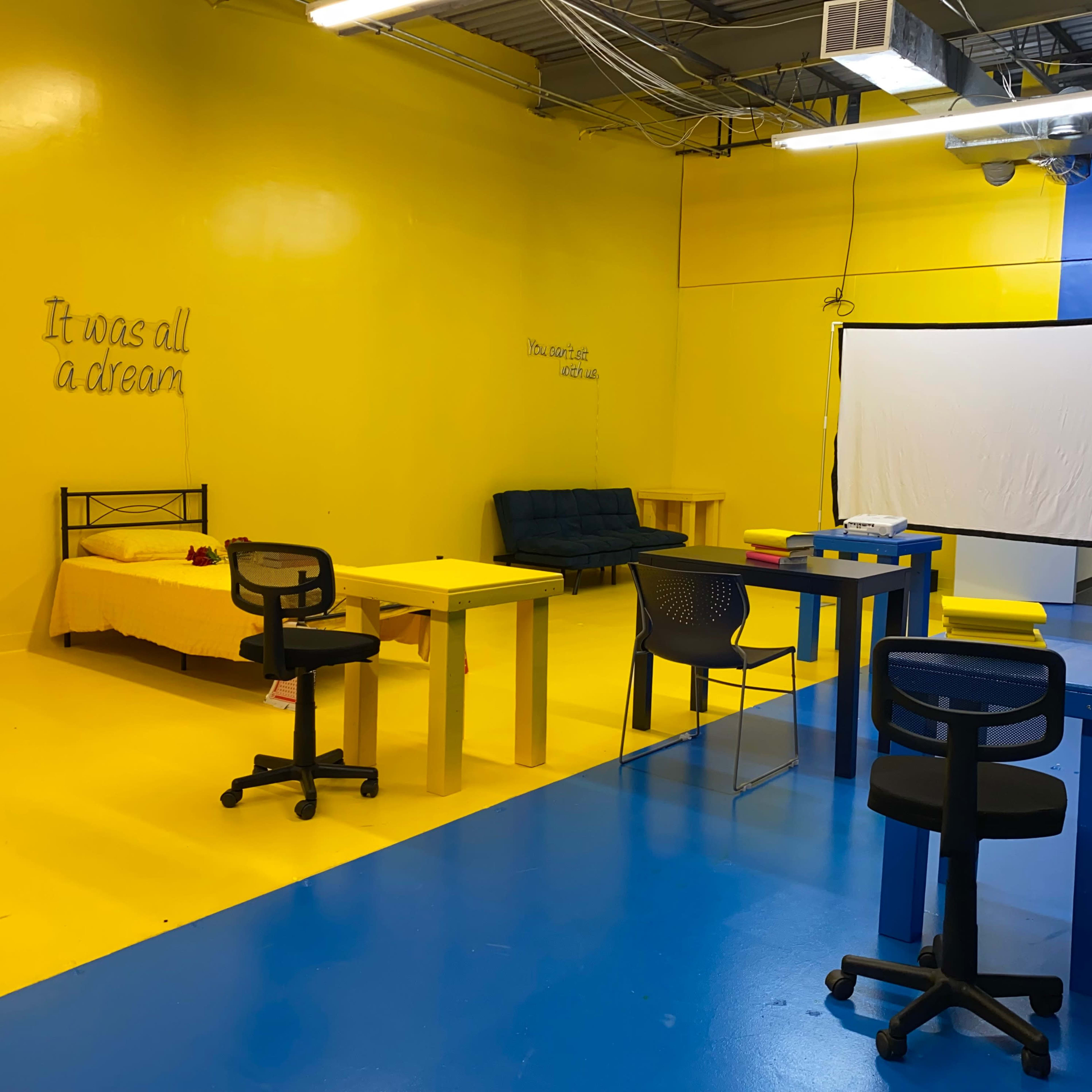 A brightly colored room with yellow walls and blue flooring, furnished with several tables and chairs, and a projection screen at one end.