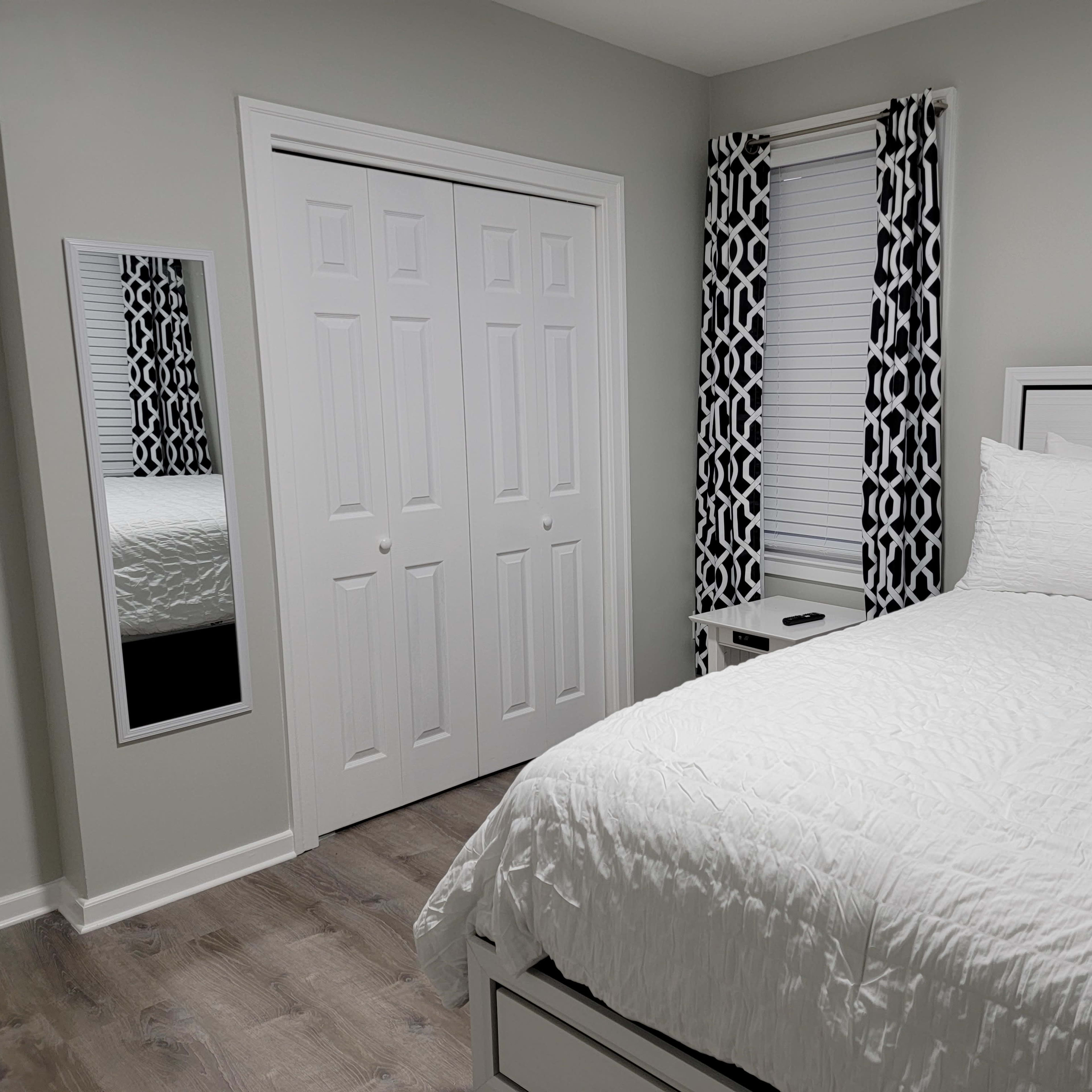 A neatly arranged bedroom featuring a white bed with decorative pillows, a mirrored closet door, and patterned curtains.